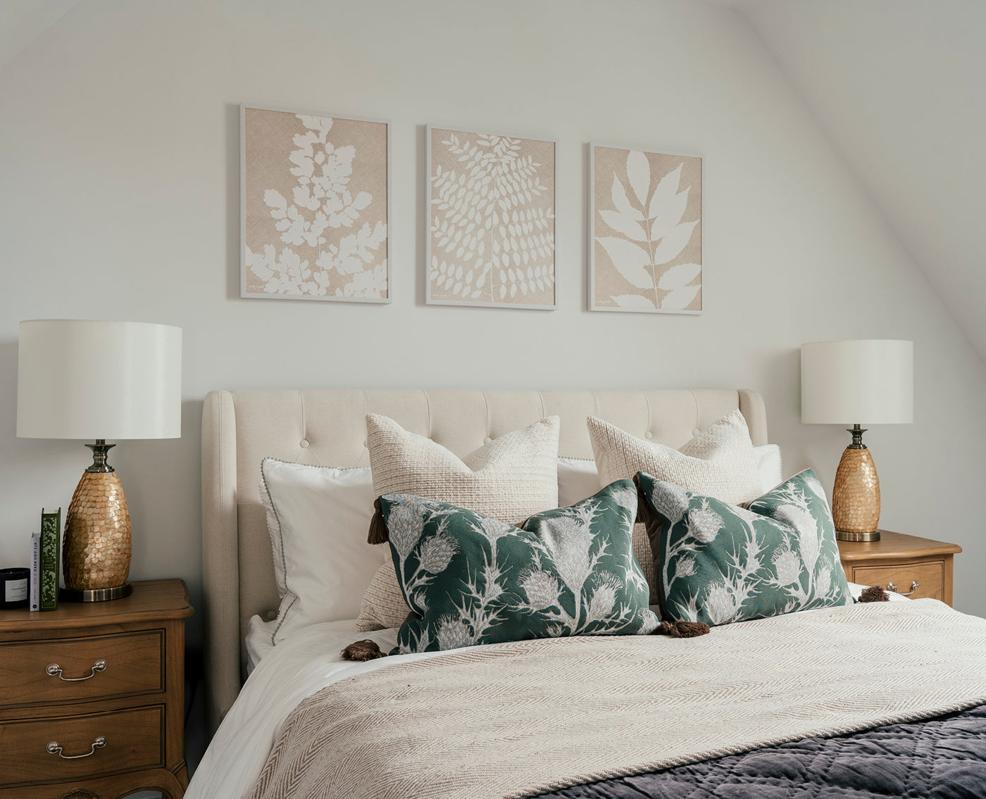 A serene bedroom with a tufted beige headboard, flanked by matching wooden nightstands and gold-textured table lamps with white shades. The bed is styled with white and textured beige pillows, complemented by two green botanical-print cushions. Above the bed, a series of four minimalist botanical prints in neutral tones decorates the wall.