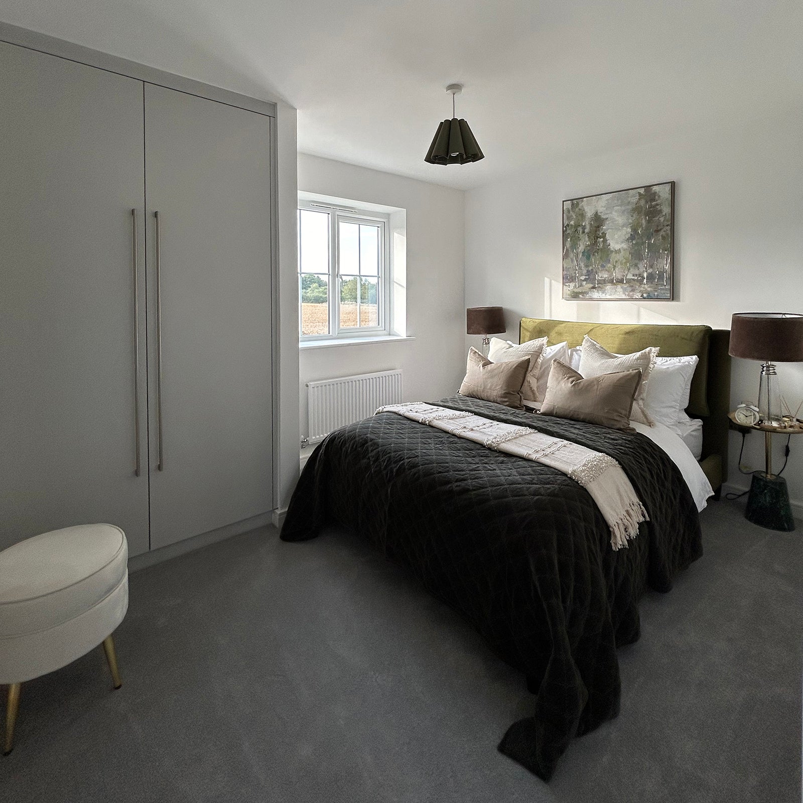 Contemporary bedroom with an olive green headboard, dark quilted bedspread, and neutral-toned cushions. Built-in wardrobes, matching bedside tables with brown lamps, and a nature-themed painting create a calm, modern space with soft grey carpet underfoot.
