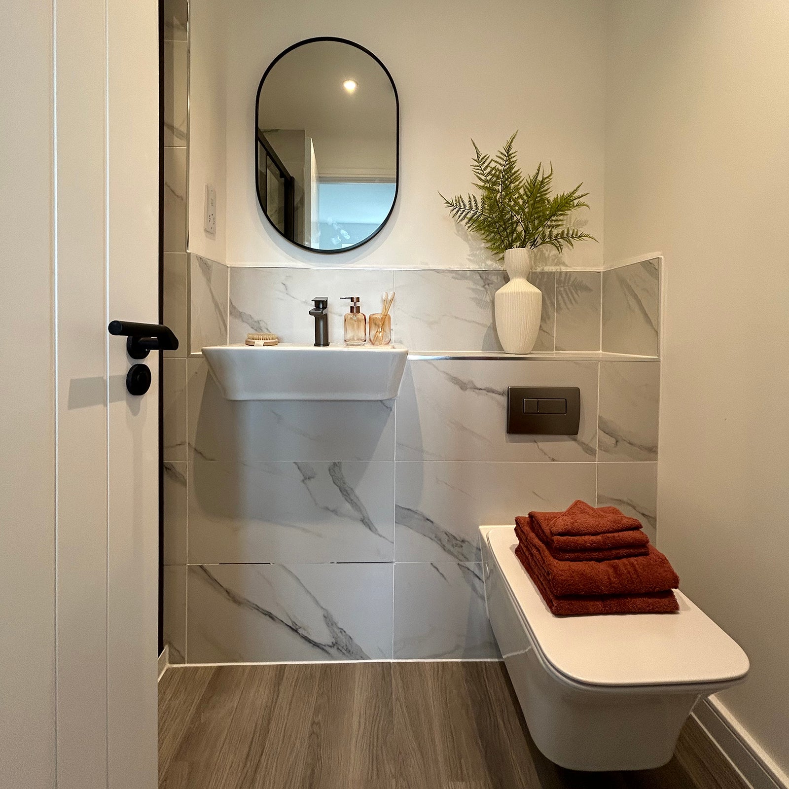 Modern cloakroom with marble-effect tiles, a floating white sink, and wall-mounted toilet. Warm accents include rust-coloured towels and a decorative fern in a textured white vase, adding a natural touch.