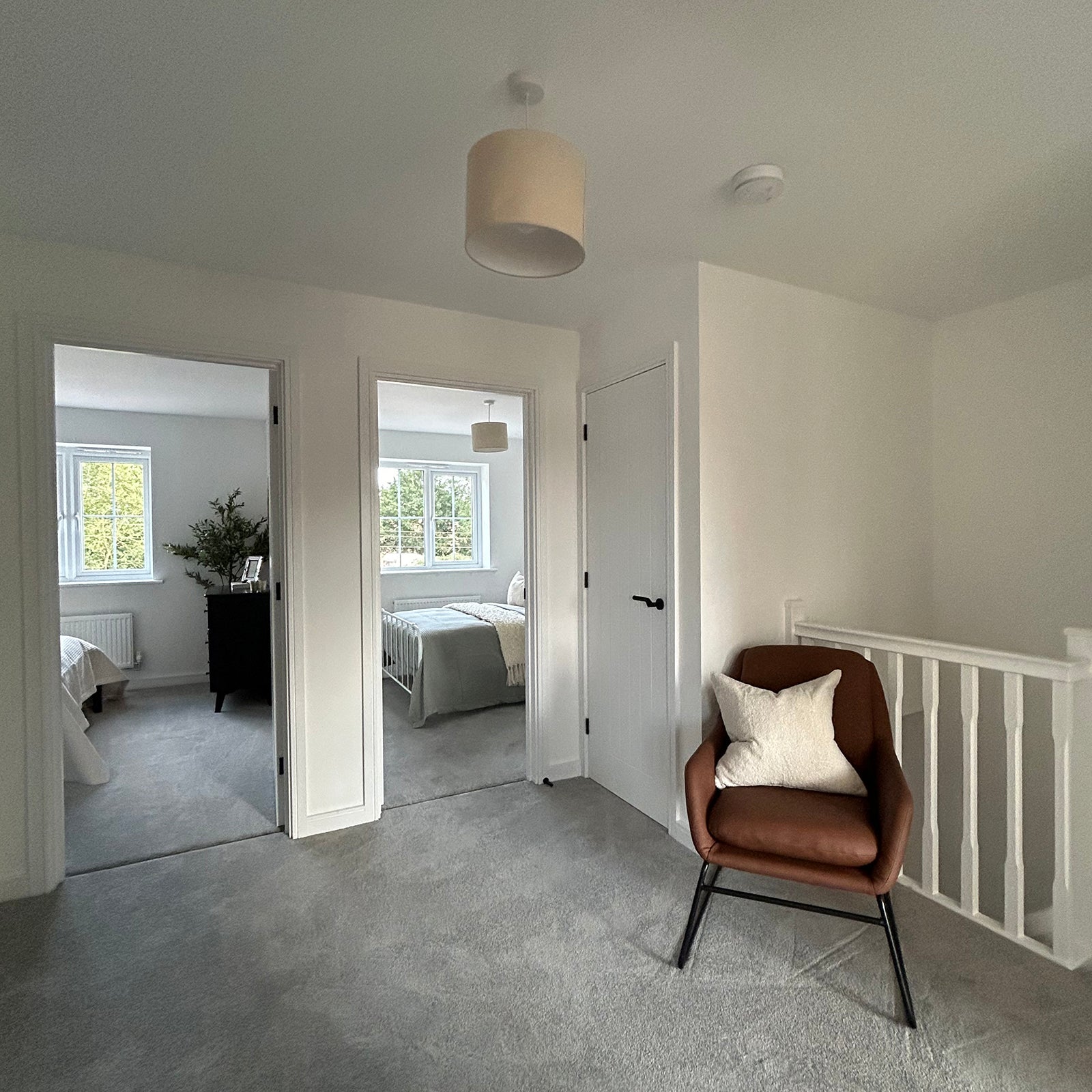 Bright landing area with light grey carpet, featuring a brown accent chair with a cream cushion. Two bedrooms are visible through open doorways, both with large windows letting in natural light.