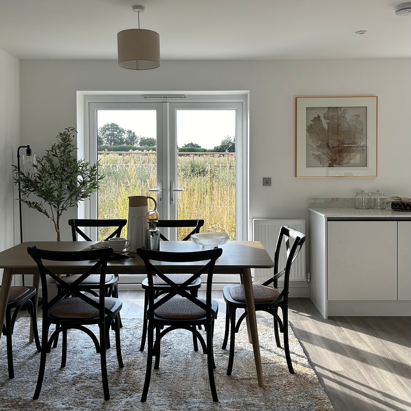 Modern dining area with a wooden table and six black chairs, set on a light rug. French doors open to a field view, allowing natural light to brighten the room.