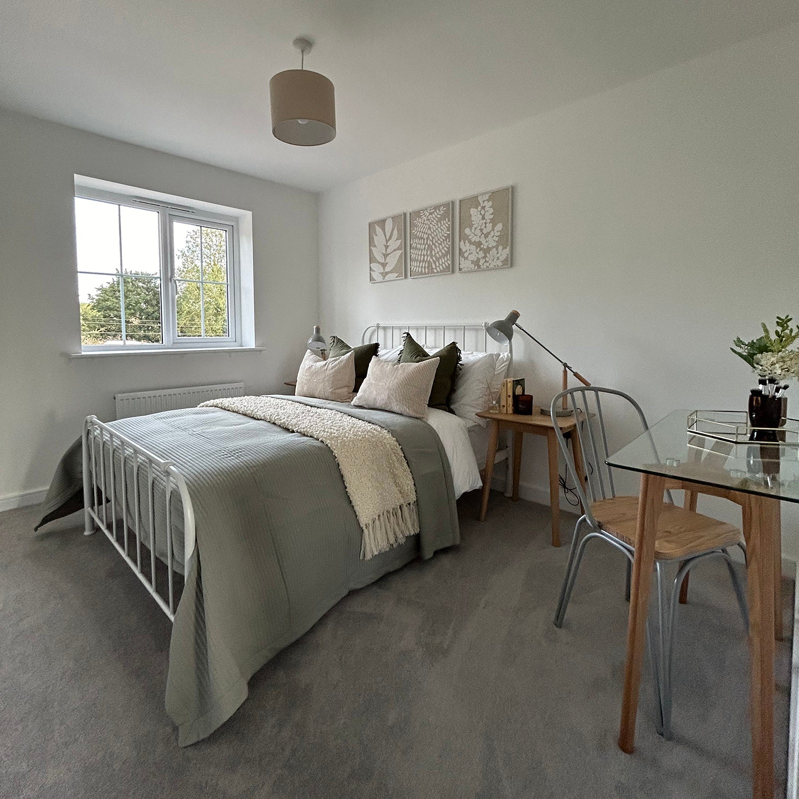 Bright bedroom with a white metal bed frame, green and neutral bedding, a small wooden desk with a reading lamp, and a glass-topped table with metal and wood chairs near the corner.