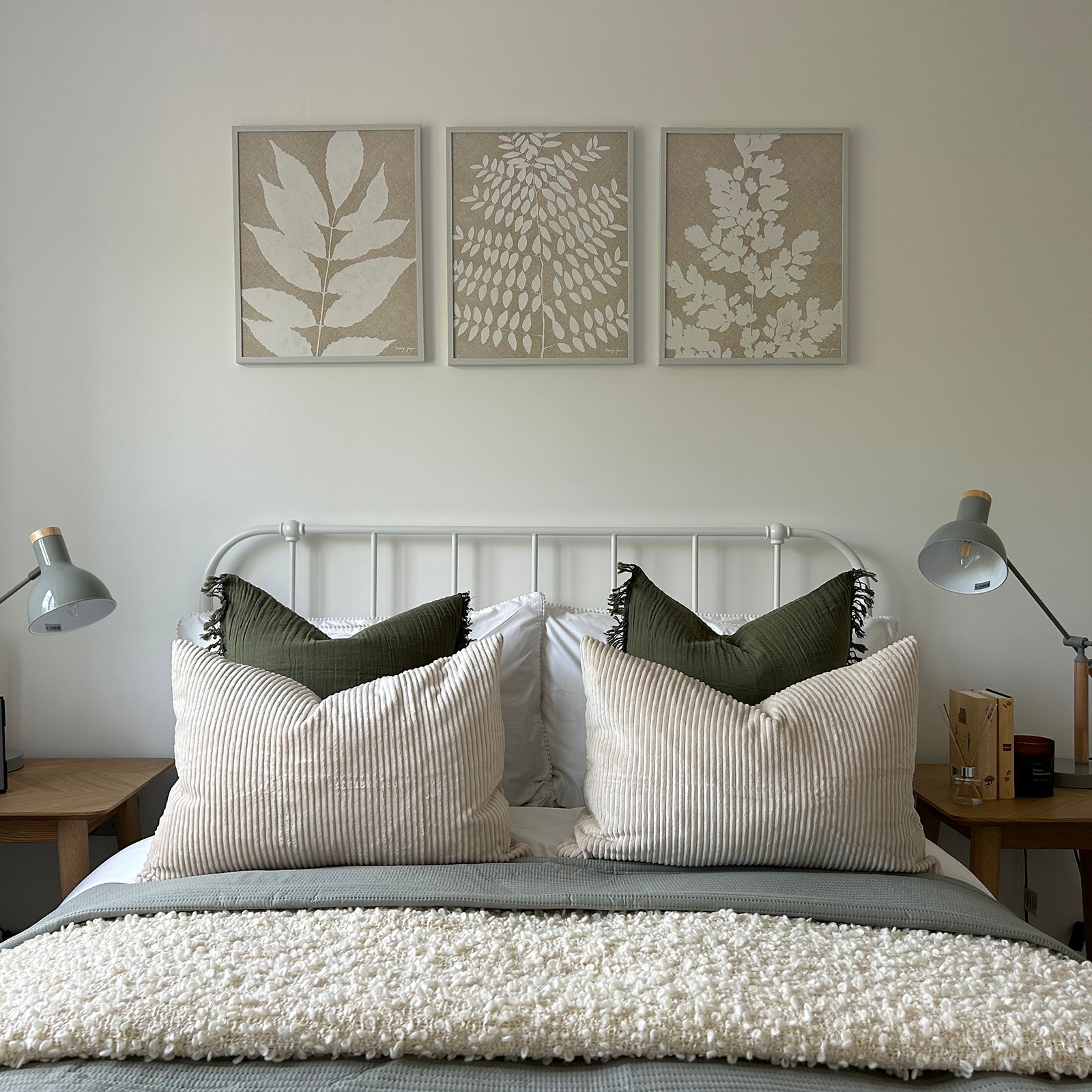 Close-up of a neatly made bed with a white metal frame, featuring layered pillows in cream and green, a textured throw blanket, matching bedside tables with grey lamps, and three botanical prints above the headboard.