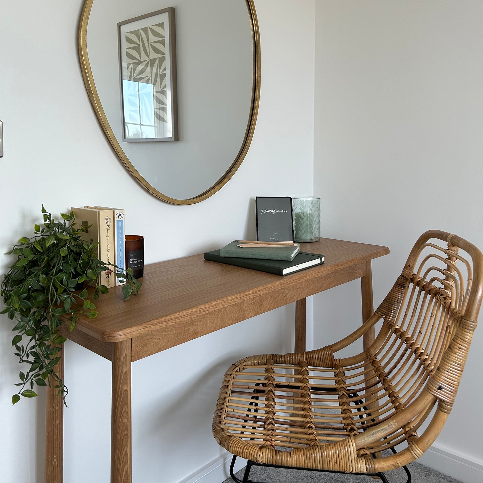 A minimalist home workspace featuring a wooden desk with a rattan chair, decorated with a trailing plant, books, a candle, notebooks, and a framed quote, all set against a white wall with a large round mirror above.