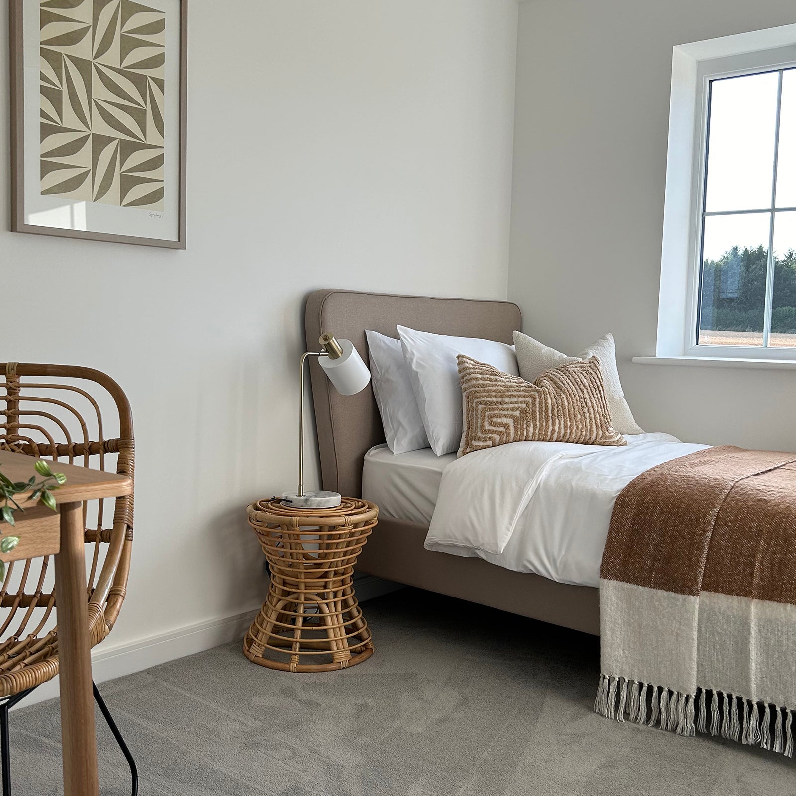 A cozy and stylish bedroom corner featuring a beige upholstered bed with white bedding and textured brown cushions, a small rattan side table with a modern white lamp, and a nearby desk with a matching rattan chair, all set against white walls and a large window letting in natural light.