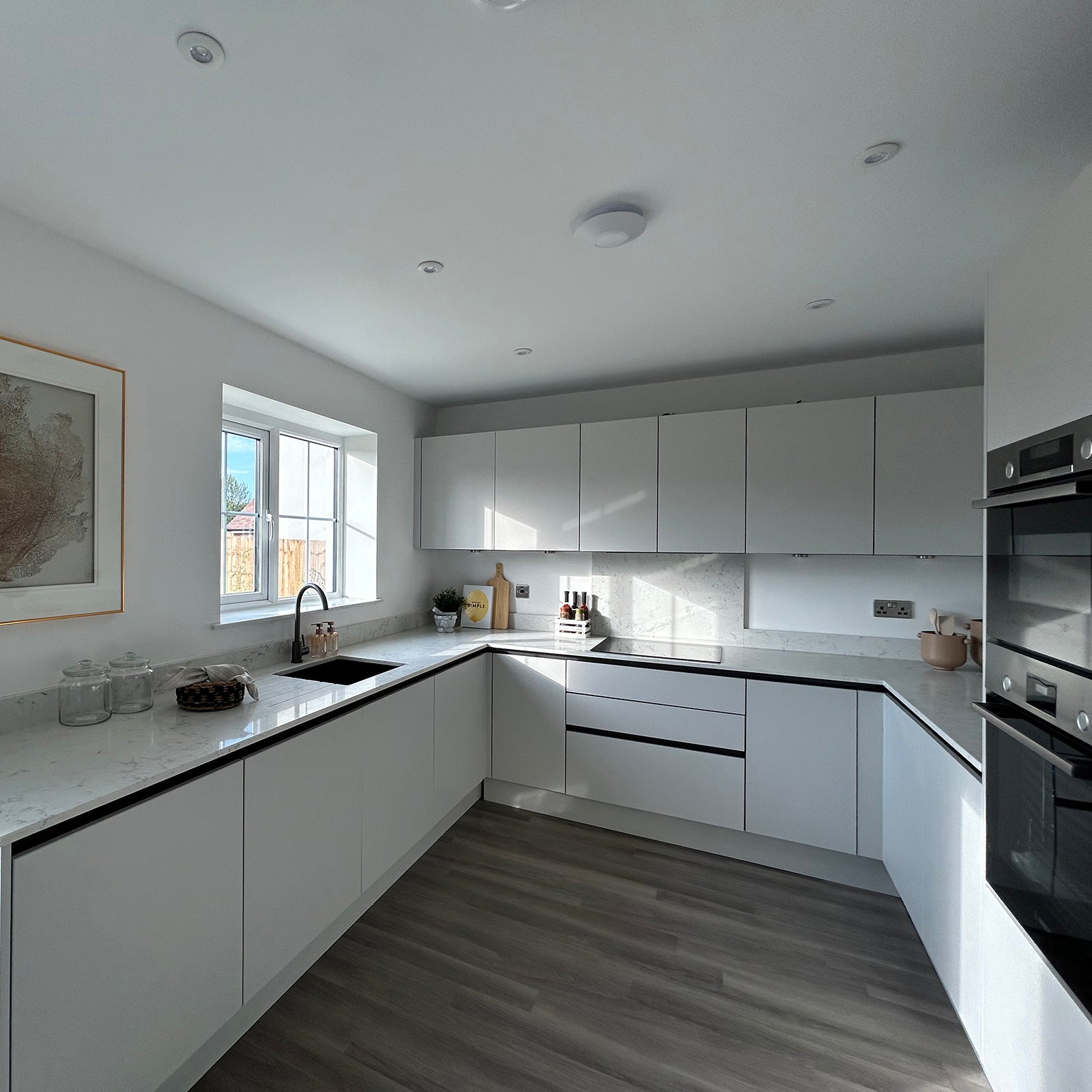 A modern, minimalist kitchen with sleek white cabinetry, integrated appliances, and a U-shaped layout featuring marble-effect countertops, under-cabinet lighting, and a large window that fills the space with natural light.
