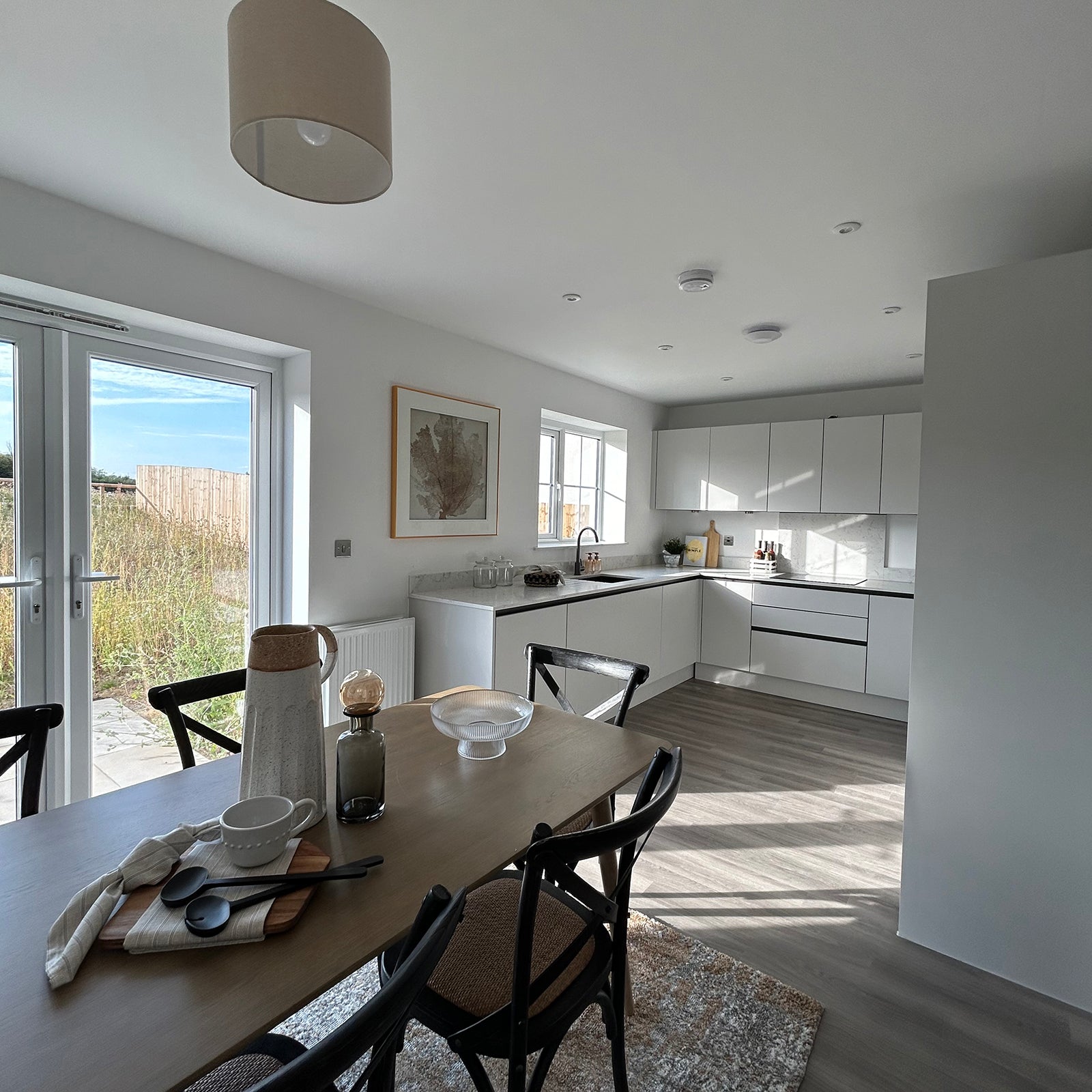 A bright, open-plan kitchen and dining area with modern white cabinetry, a wood dining table set for four, large glass doors leading to a garden, and natural light streaming in through multiple windows, creating a warm and inviting atmosphere.