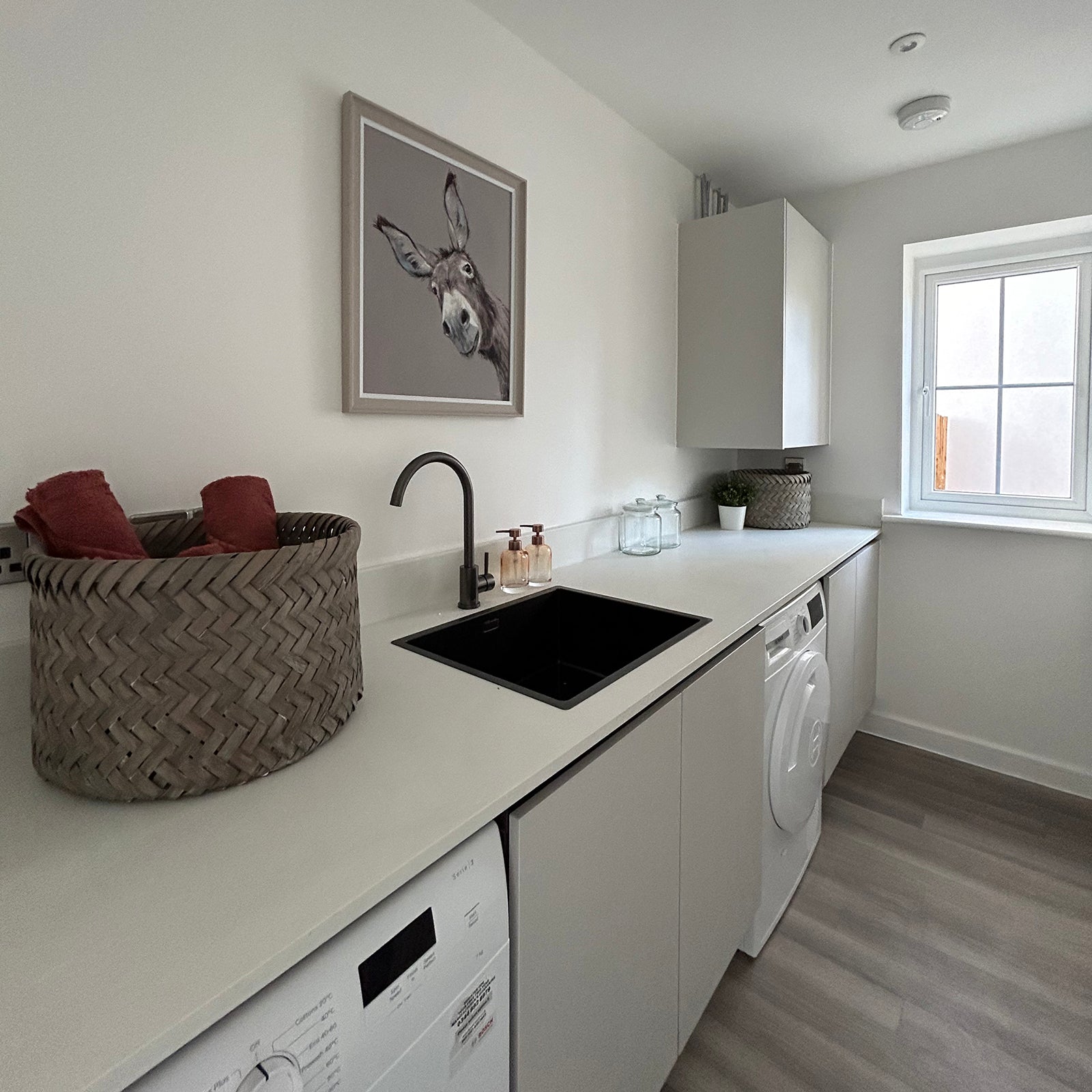A modern utility room featuring sleek white cabinetry, a black sink with a matching faucet, a washing machine, and a wicker basket with red towels; a playful framed picture of a donkey adds character above the sink, and natural light enters through a window at the far end.