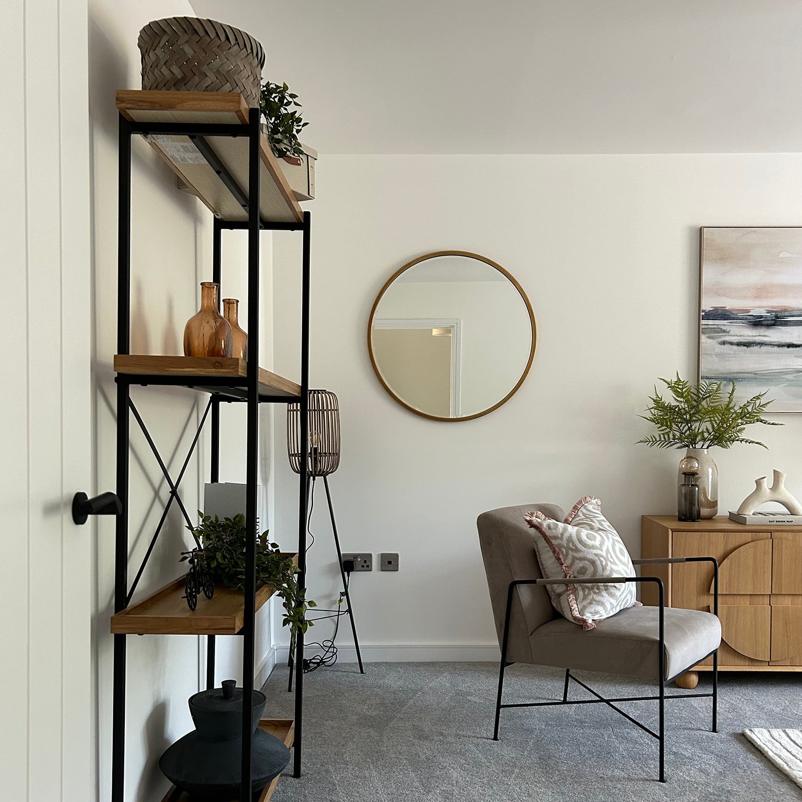 A modern living space with an industrial-style shelf, a neutral armchair with a patterned cushion, a wooden sideboard, and decorative touches like plants, a round mirror, and abstract artwork.
