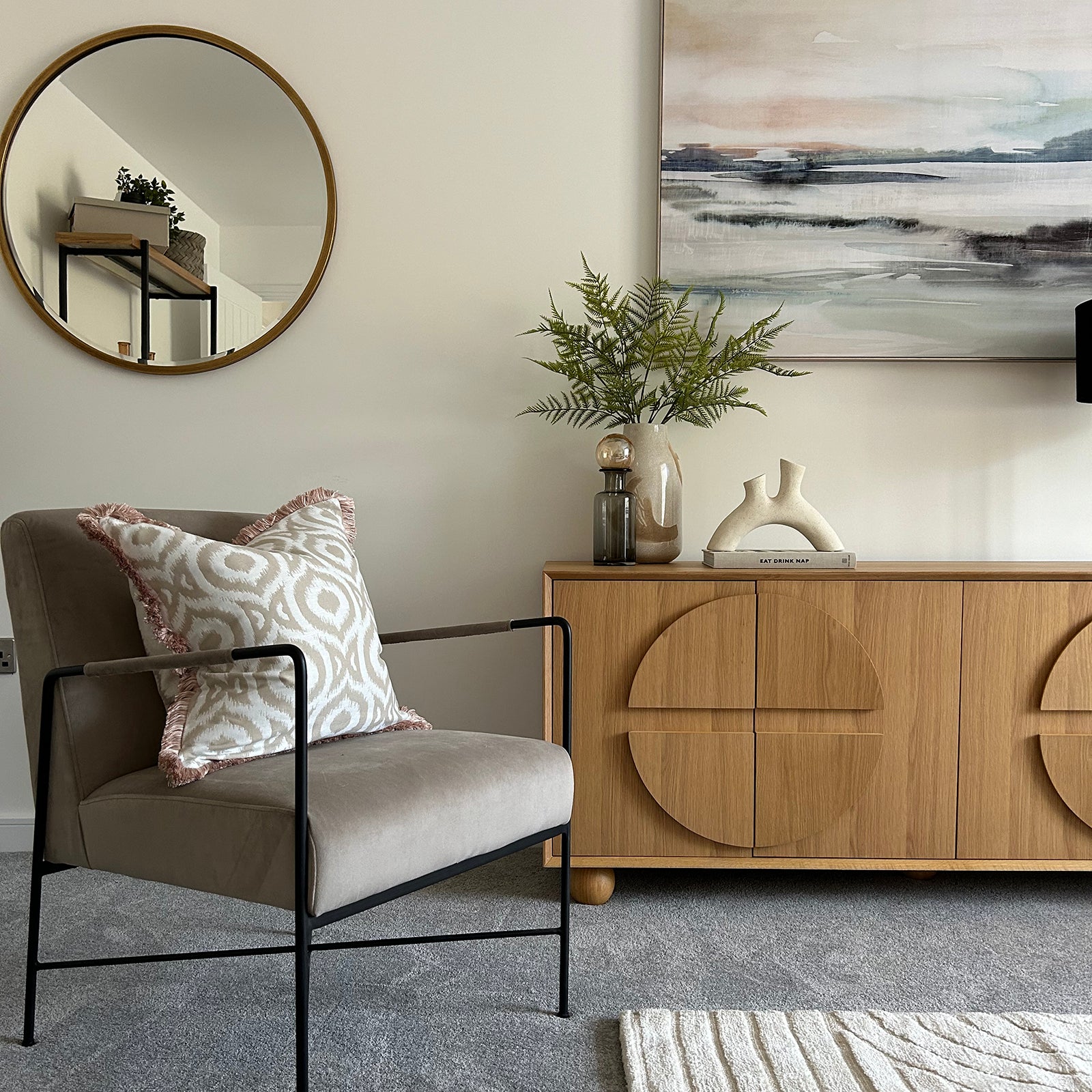 Close-up of a stylish living room corner with a modern armchair, patterned cushion, circular mirror, and a wooden sideboard decorated with vases, books, and greenery.