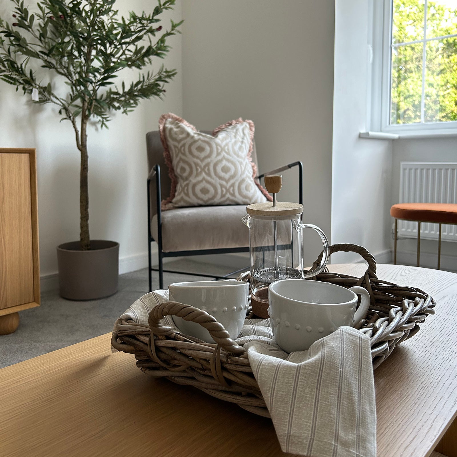 Close-up of a coffee table with a wicker tray holding two white mugs, a French press, and a striped napkin, with a cozy chair and potted tree in the background.