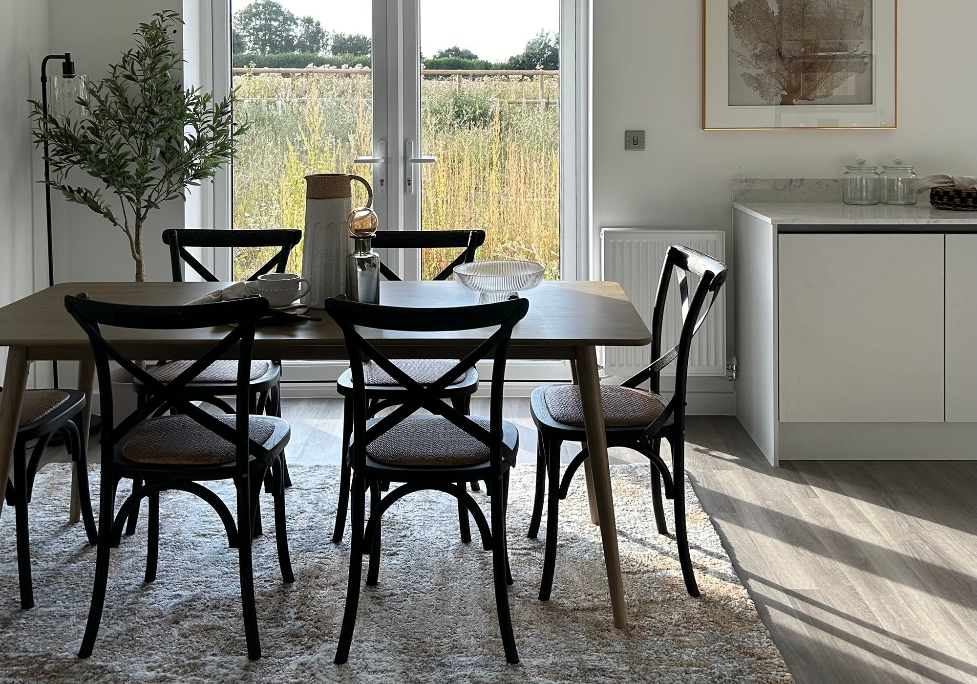 Modern dining area with a wooden table surrounded by black cross-back chairs on a soft rug, set against tall glass doors opening to a sunlit field, with minimalist kitchen cabinetry and decor nearby.