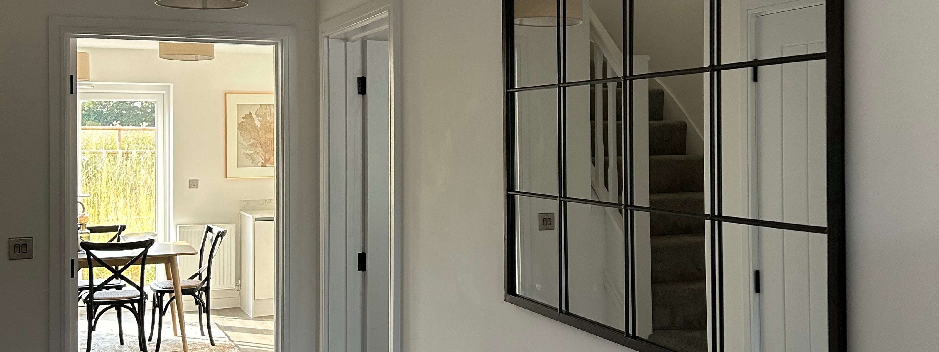 Bright hallway featuring a large grid-style mirror reflecting a staircase, with a view into a sunlit dining area furnished with a wooden table, black cross-back chairs, and a framed artwork on the wall.
