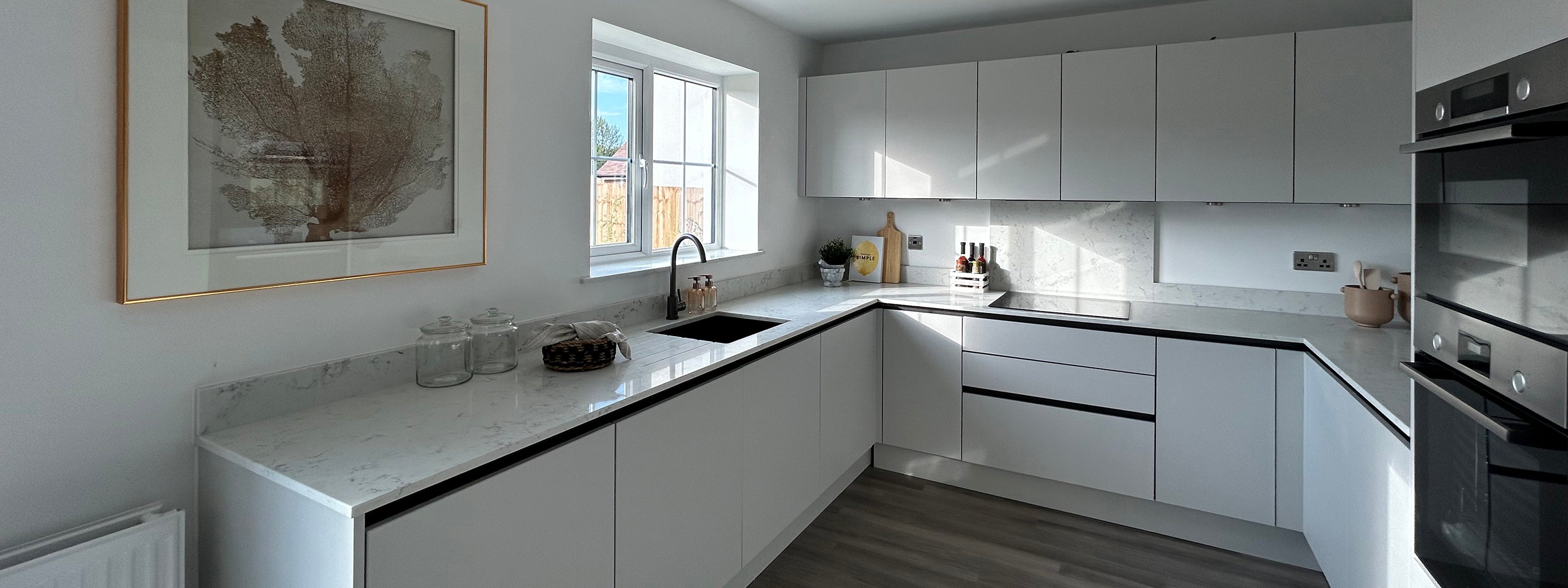 Modern kitchen with sleek white cabinets and marble countertops, featuring a large window above the sink, minimalist decor including a framed botanical print, and a mix of functional and decorative items on the counters.