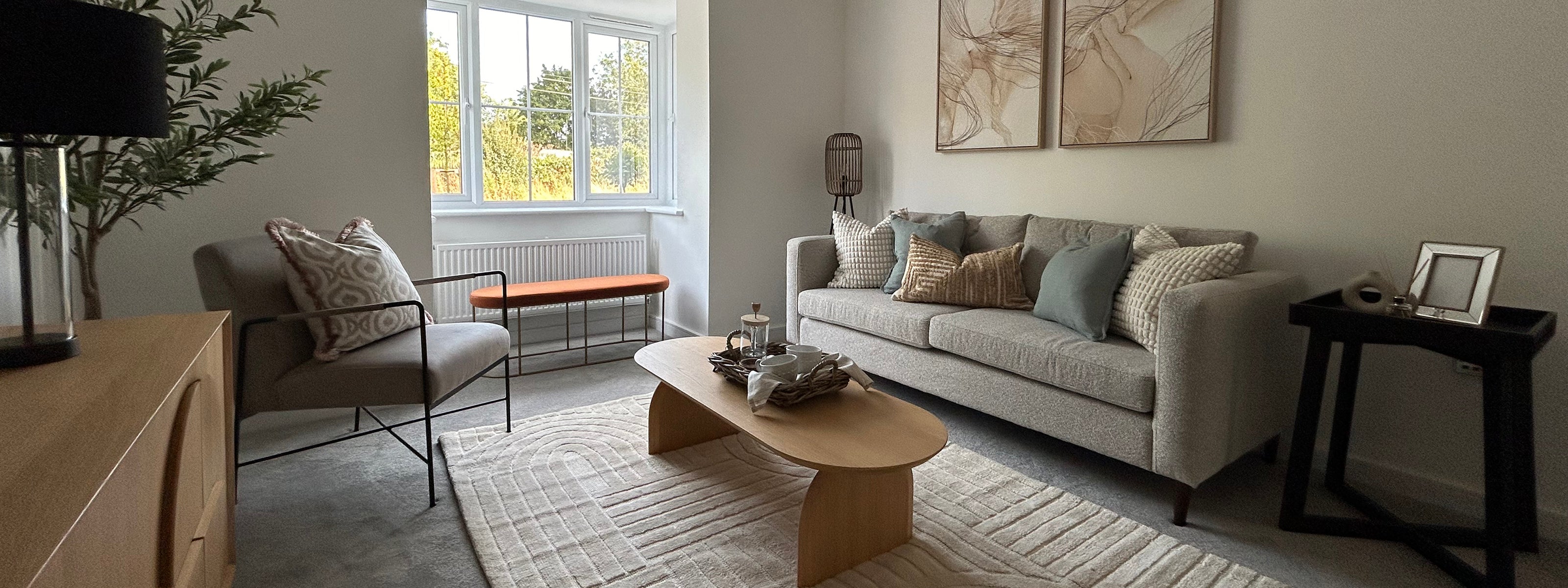 Contemporary living room with a large window allowing natural light to fill the space, featuring a neutral-toned sofa with assorted throw pillows, abstract wall art, a light wood coffee table on a textured rug, and minimalistic decor including a bench, indoor plant, and modern lighting.
