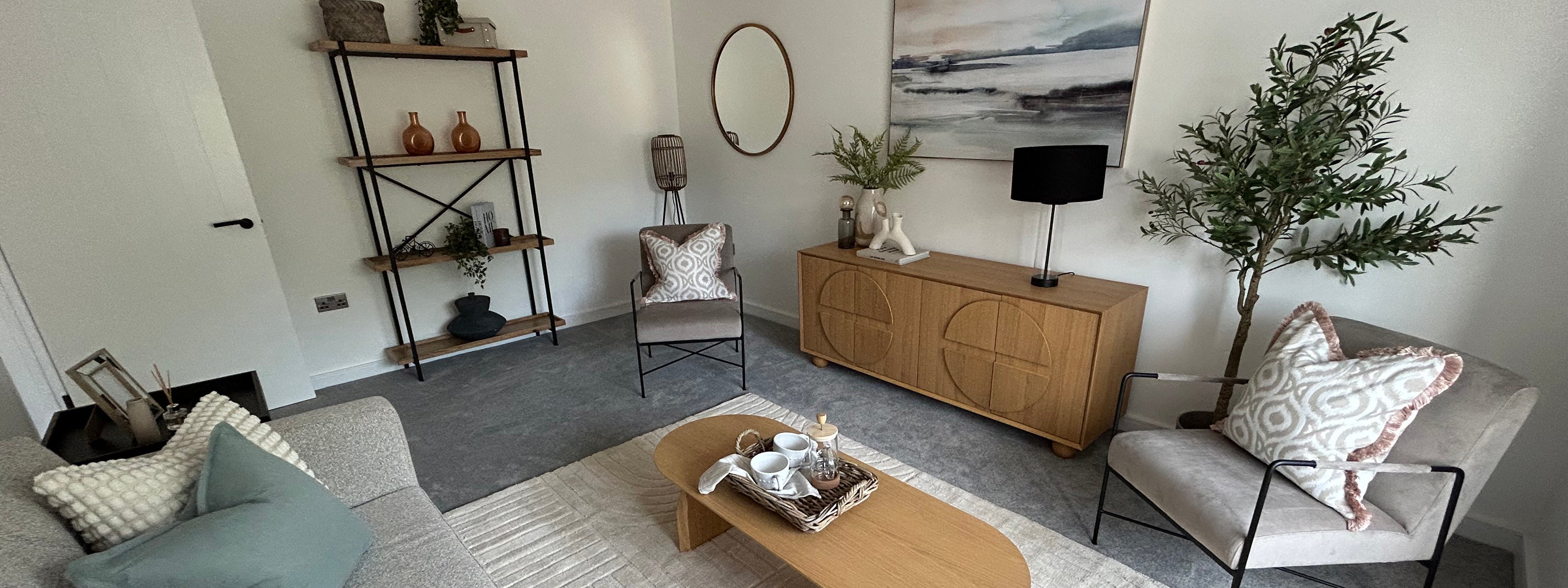 Modern living room with neutral tones and minimalist decor, featuring a beige sofa and accent chairs, a light wooden coffee table with a tray of tea items, a wooden sideboard with geometric design, wall-mounted artwork, a round mirror, open metal shelving with decorative items, and indoor plants for a natural touch.