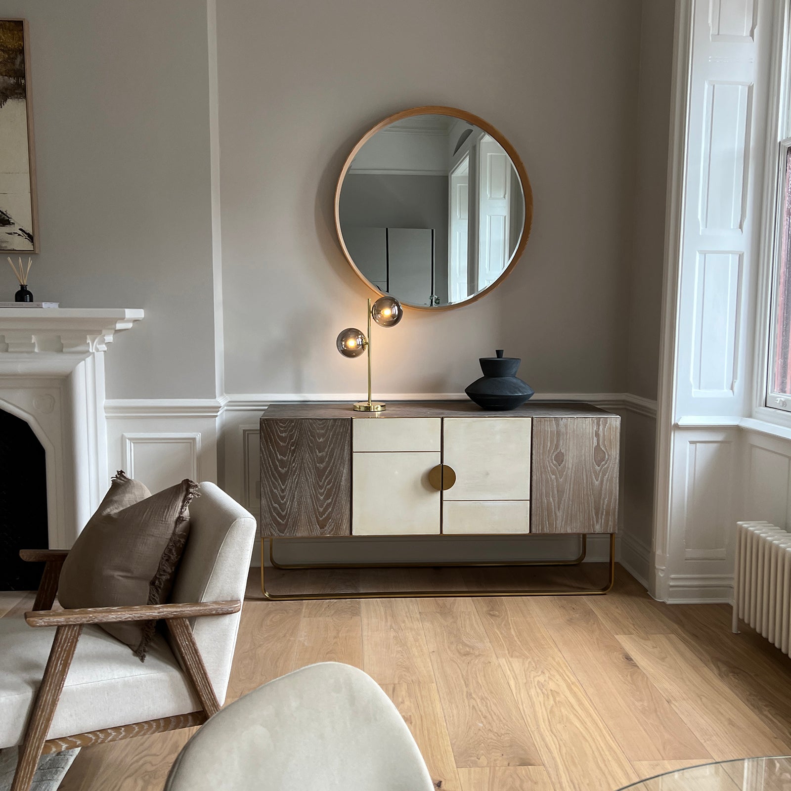Elegant living room corner featuring a mid-century modern sideboard with a mix of wood and cream-toned panels, a round wooden-framed mirror above, a sculptural black vase, and a stylish brass lamp with two globe bulbs, all set against light grey walls with white trim and natural wood flooring.
