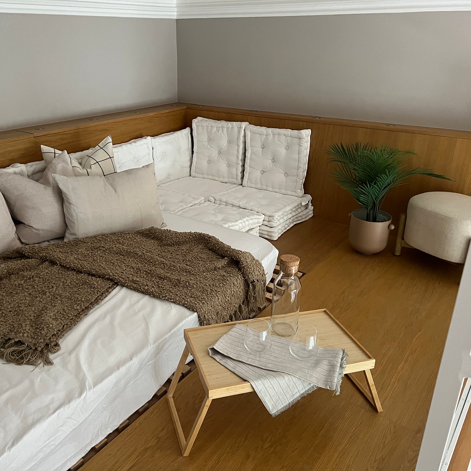 Cozy corner seating area with cushioned white floor mattresses, layered neutral pillows, a brown throw blanket, and a light wood tray table with glassware, set against warm wood paneling and accented by a potted plant.