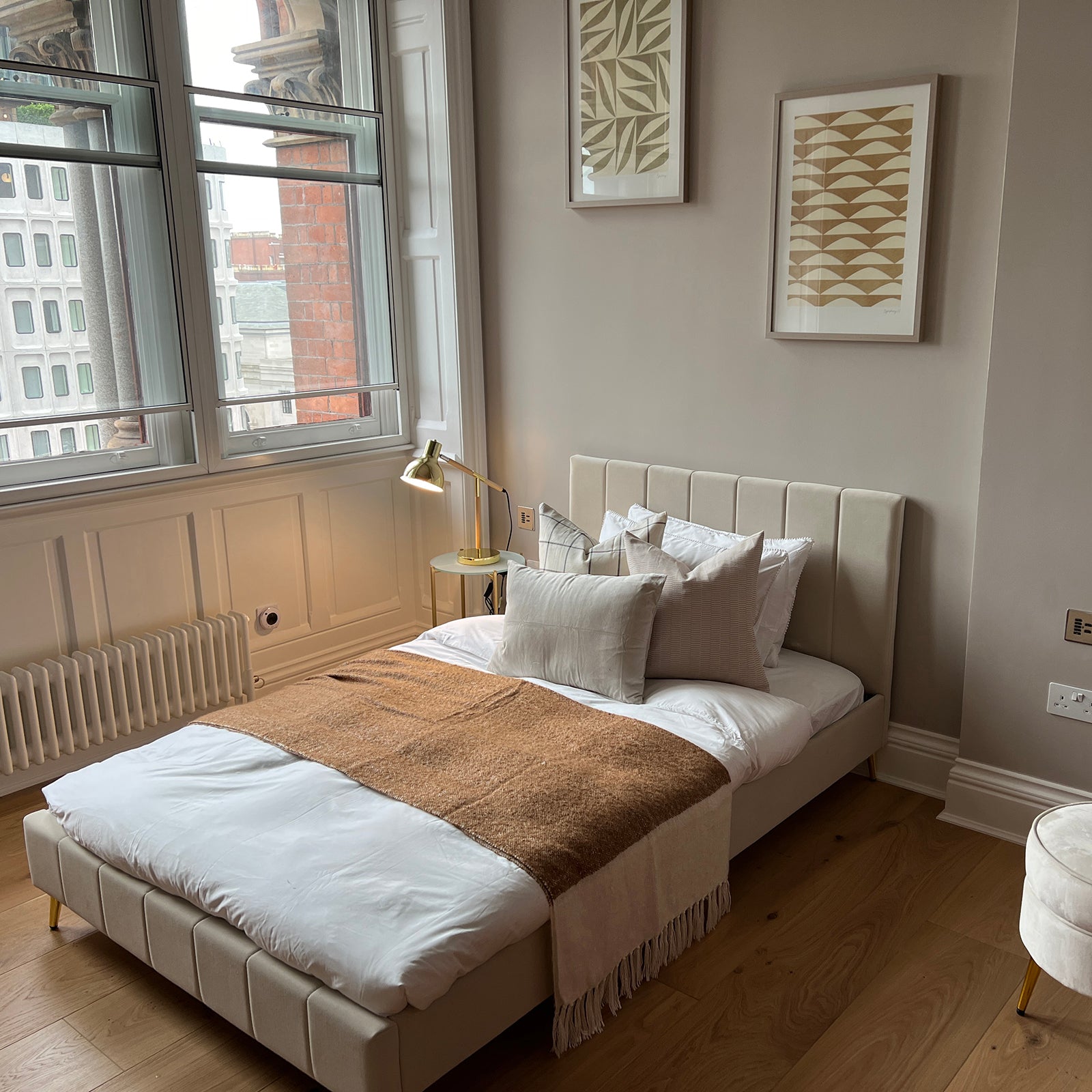 Bright, minimalist bedroom featuring a low-profile upholstered bed with layered neutral pillows, a tan throw blanket, gold side lamp, abstract wall art, and large windows with classic white trim overlooking a city street.