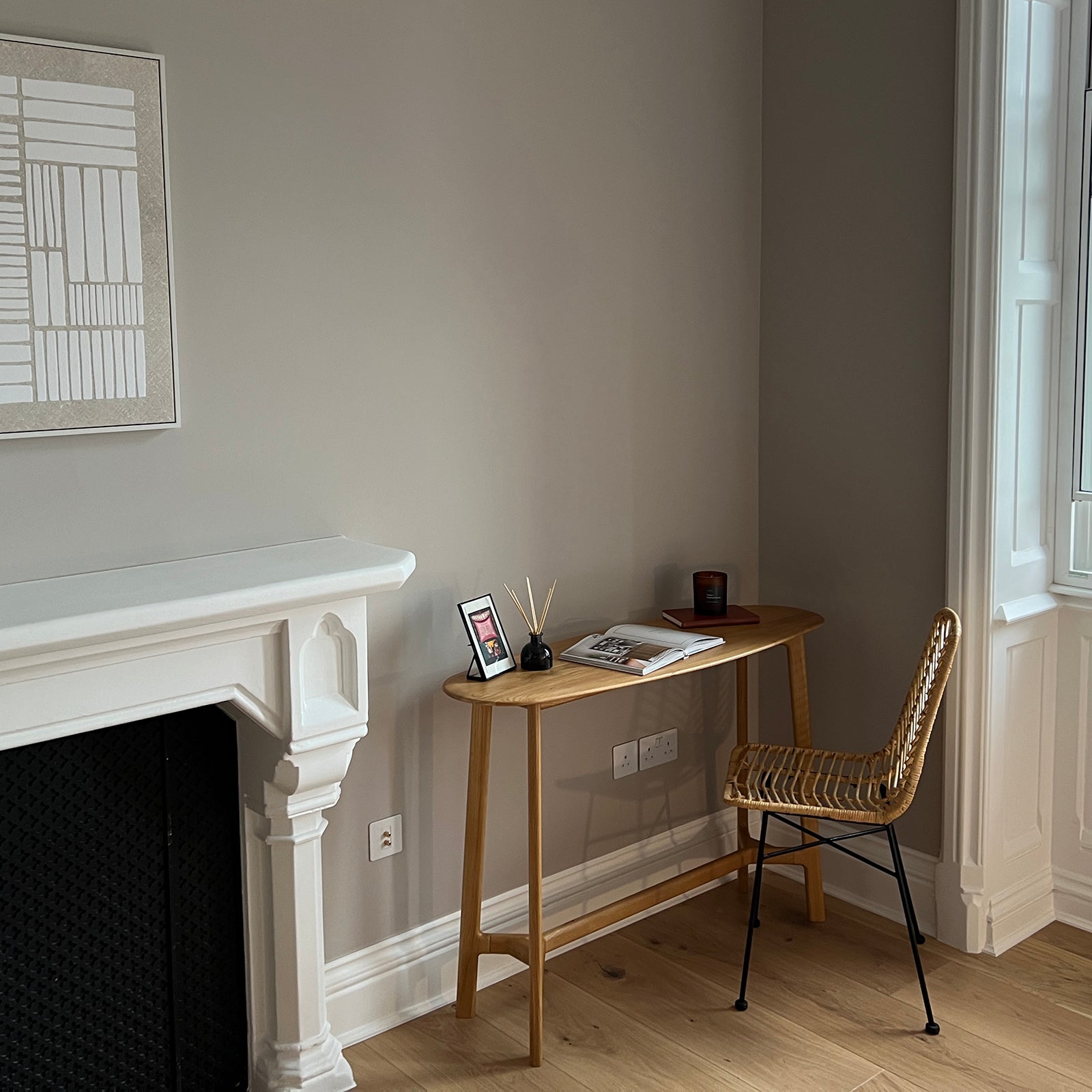 A small, stylish workspace featuring a light wooden desk with minimalist decor, including a reed diffuser, candle, and open book, paired with a woven rattan chair on black metal legs, next to a traditional white fireplace and large window with classic moulding.