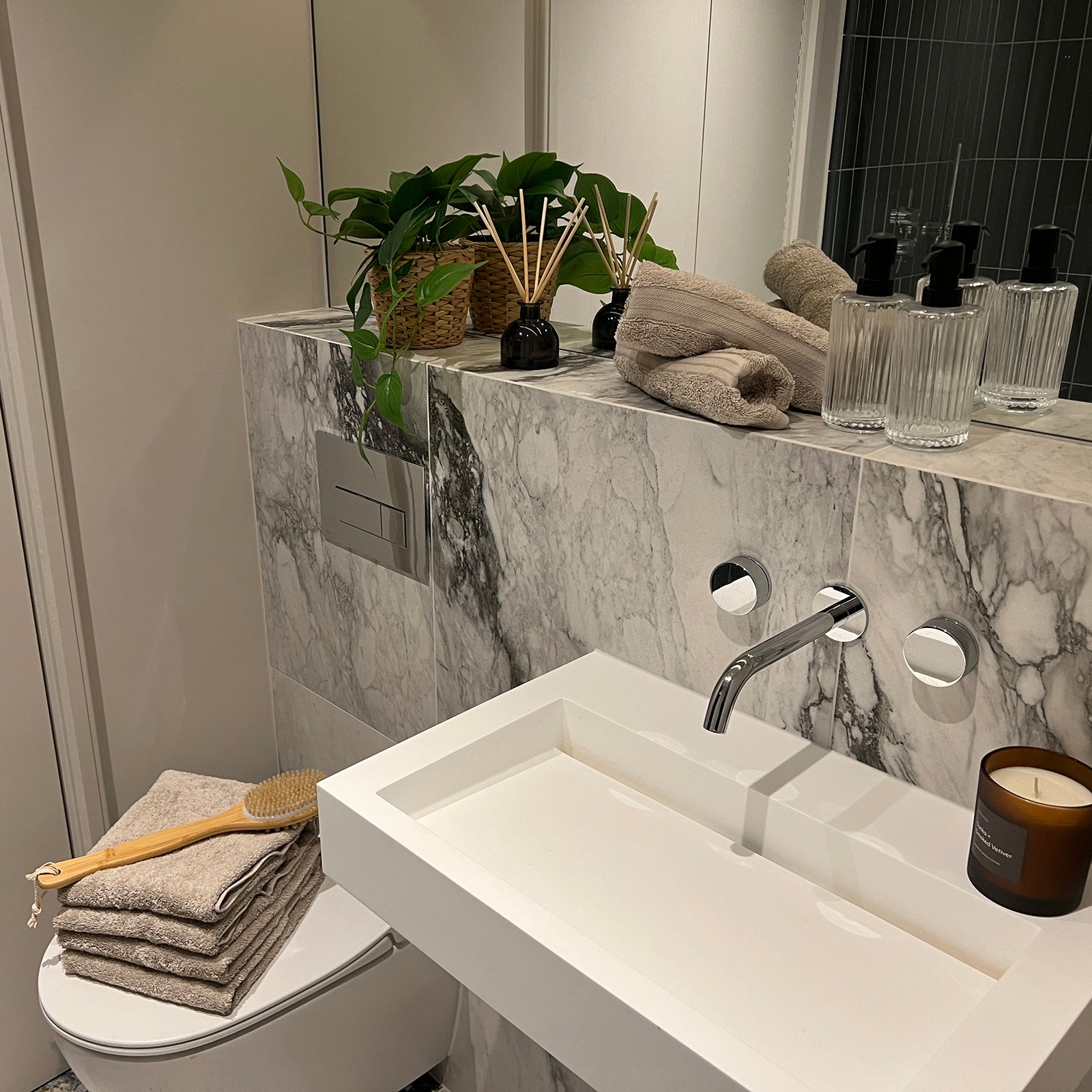 Modern bathroom vanity with marble detailing, featuring a white rectangular sink with a wall-mounted chrome tap, a back shelf styled with soft taupe towels, ribbed soap dispensers, reed diffusers, a potted plant in a wicker basket, and a lit candle; neatly folded towels and a wooden bath brush rest atop the toilet.