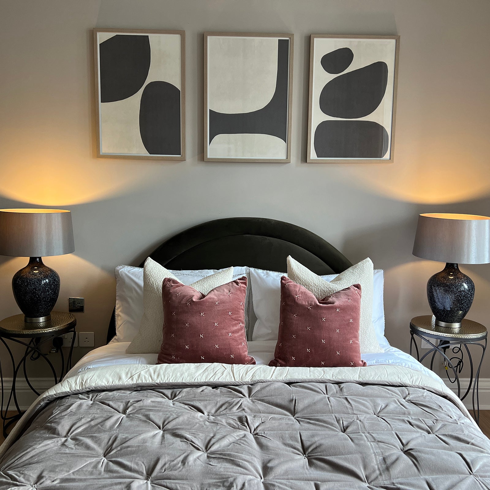 Stylish bedroom with a central double bed featuring a dark green arched headboard, dressed in white bedding, a taupe quilted blanket, and pink velvet accent cushions; flanked by two ornate black side tables with matching textured ceramic lamps, and three abstract black and cream framed artworks hung above the bed.