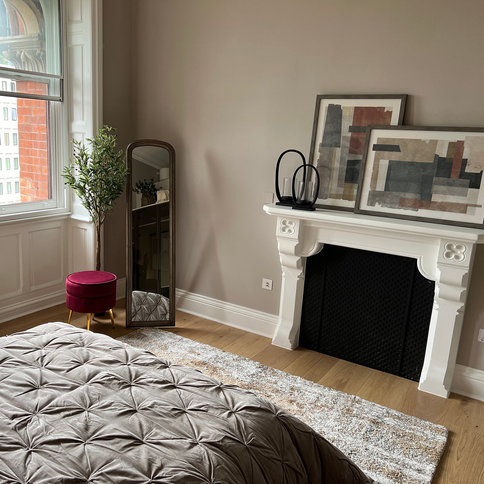 Elegant bedroom corner featuring a decorative white fireplace with two abstract framed artworks leaning against the wall above it, a tall arched mirror reflecting the bed, a faux potted tree beside the window, and a deep red velvet pouffe with gold legs; the wooden floor is partially covered by a textured area rug and the foot of a taupe quilted bedspread is visible.