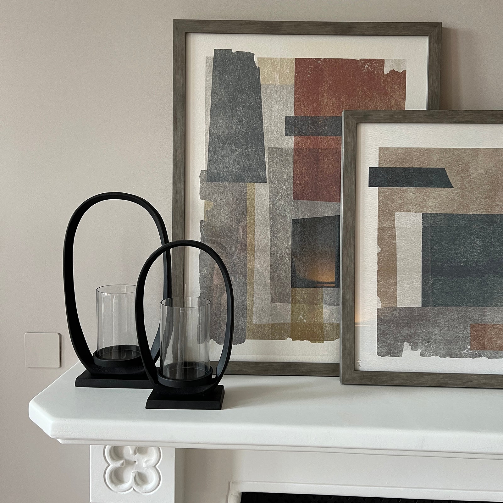 Close-up of a white decorative fireplace mantel featuring two framed abstract artworks in muted earth tones overlapping slightly, accompanied by two modern black oval candle holders with clear glass inserts, set against a light grey wall.