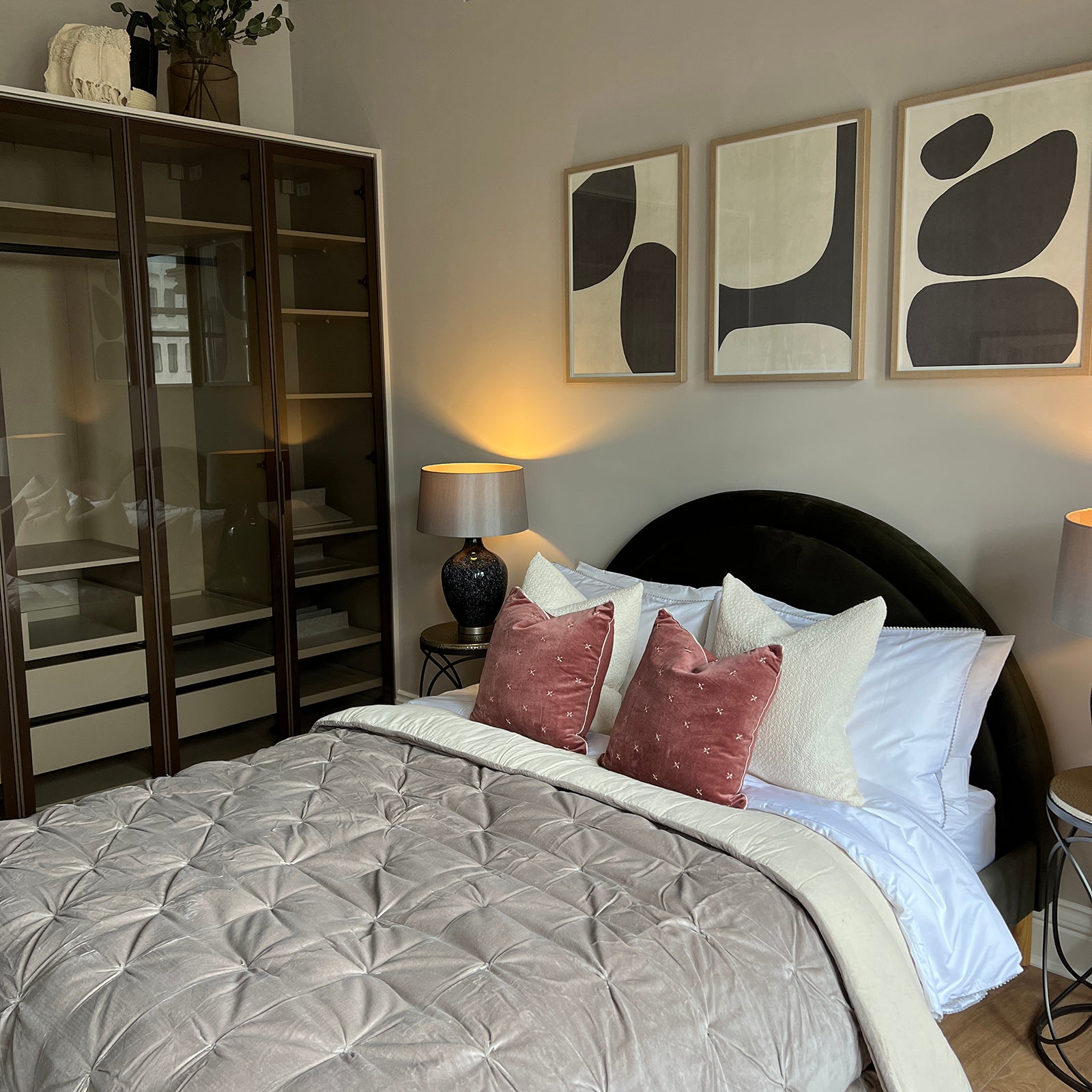 Modern bedroom with a soft, neutral colour palette featuring a neatly made bed with a tufted taupe duvet, pink and cream cushions, flanked by black ceramic lamps on side tables. Above the bed hang three abstract art prints, and to the left is a glass-fronted wardrobe with built-in shelving.