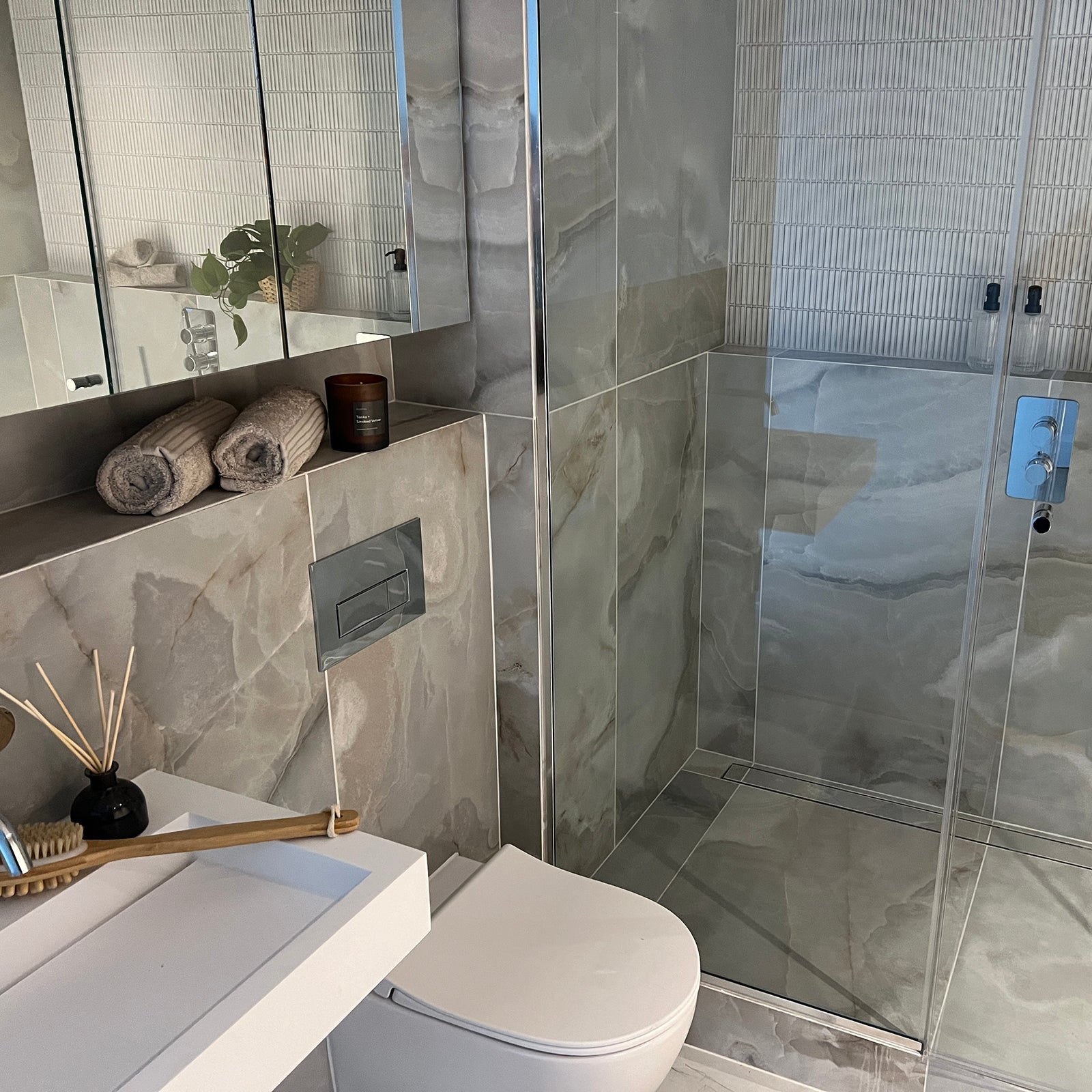 Contemporary bathroom with large marble-effect tiles, featuring a walk-in glass shower, wall-mounted toilet, and a white rectangular sink. The shelf above the toilet holds rolled beige towels, a brown candle, and a small plant, while a wooden bath brush and reed diffuser sit near the sink.