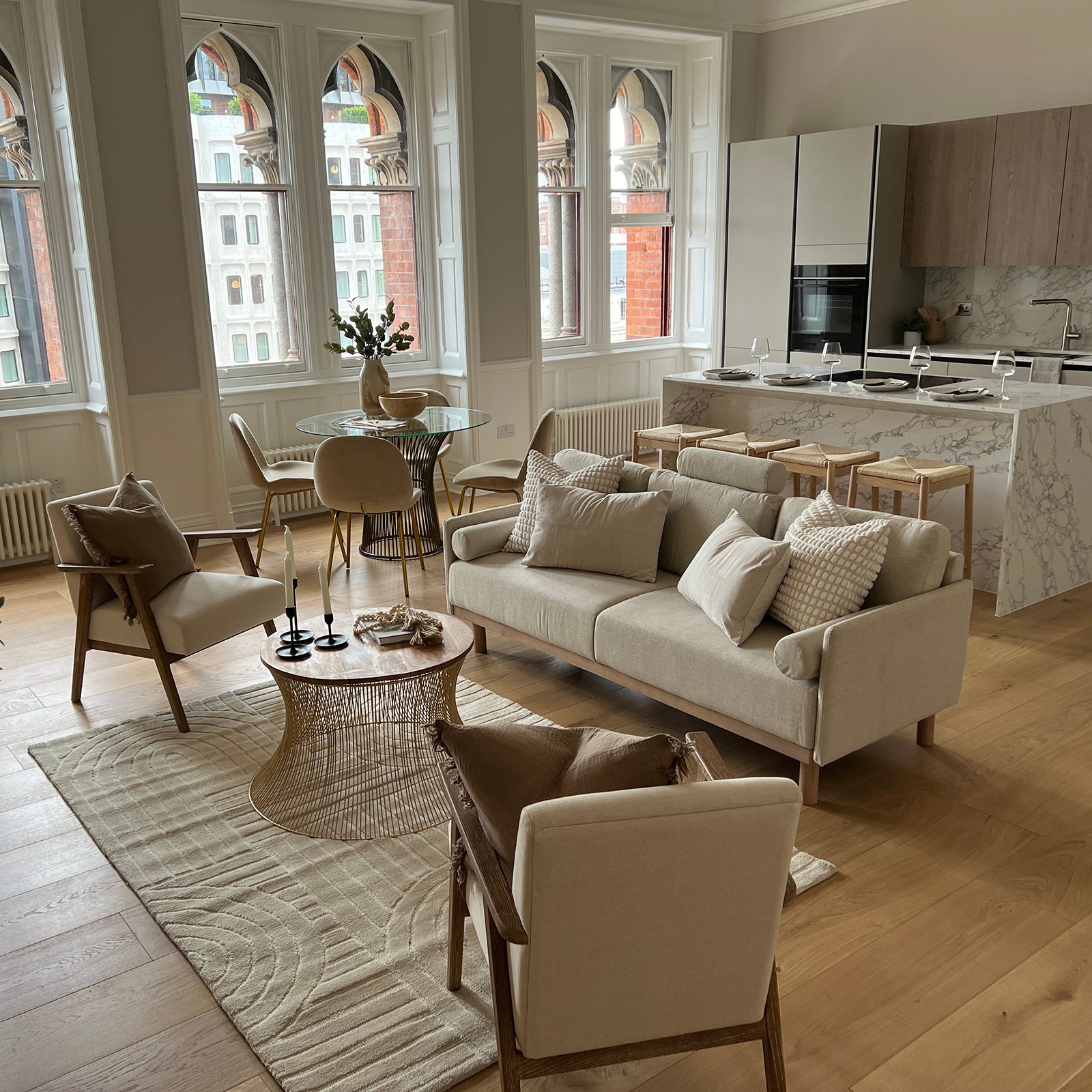 Elegant open-plan living and kitchen area with neutral-toned furnishings, including a cream sofa, wooden armchairs, a round glass dining table, and a marble kitchen island with bar stools. Large arched windows provide ample natural light and overlook a historic building facade.