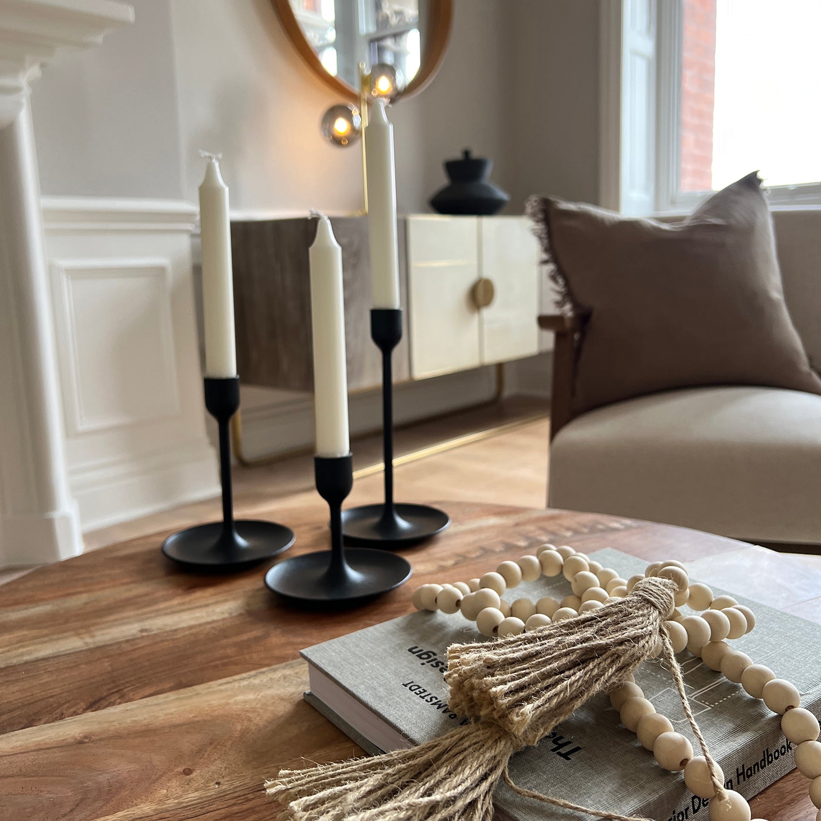 Close-up of a cozy living room scene featuring a wooden coffee table with a decorative beaded tassel, a design book, and three black candle holders with white taper candles. In the background, a beige armchair, a modern console with brass legs, and a round mirror add to the warm, minimalist aesthetic.
