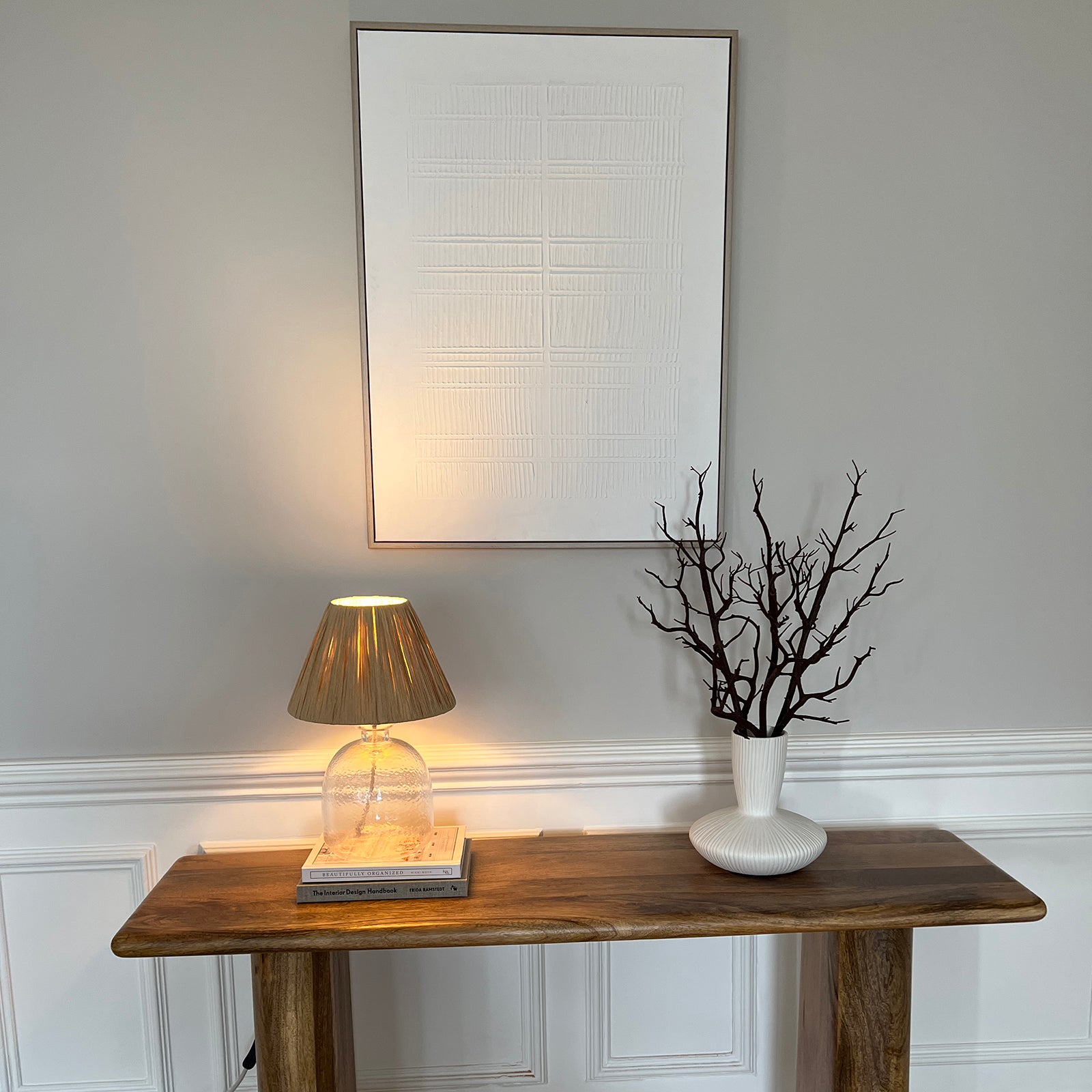 A minimalist hallway setup featuring a rustic wooden console table topped with a glass table lamp with a pleated shade, a stack of books, and a white ceramic vase holding bare black branches. Above the table hangs a large textured white artwork, all set against a soft grey wall with white wainscoting.