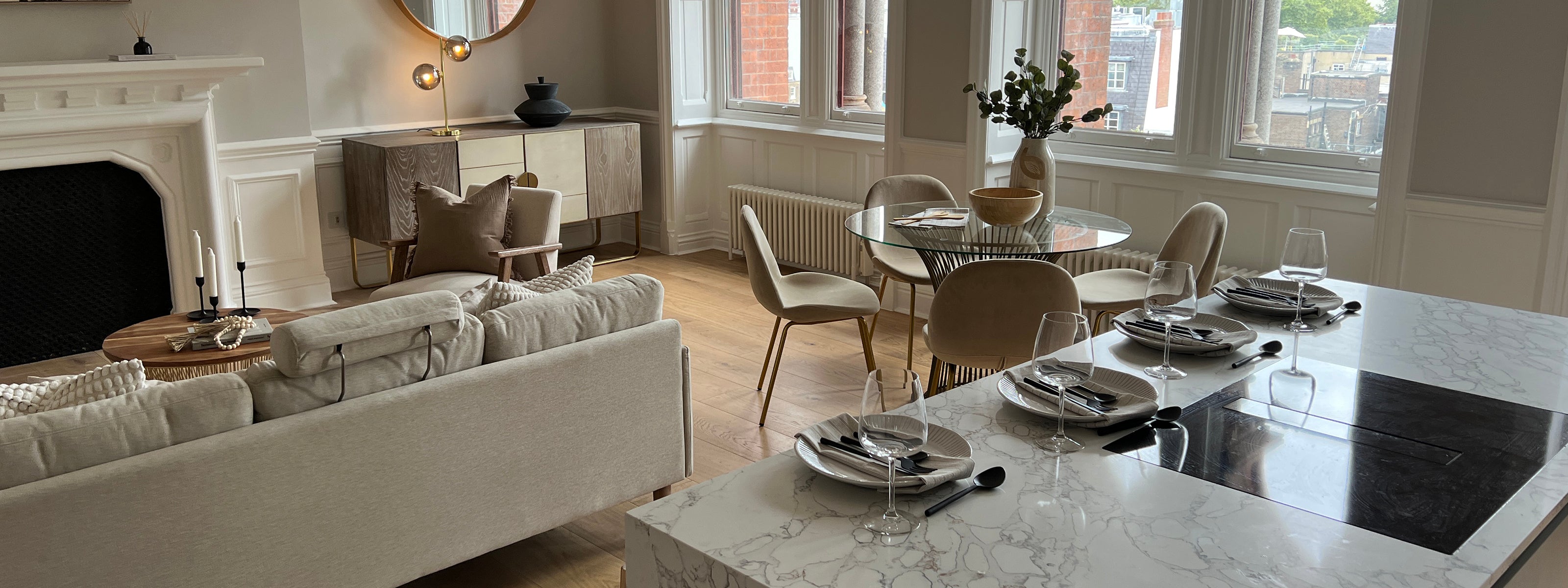Elegant open-plan interior with a marble kitchen island, beige sofa, round dining table, and large windows.