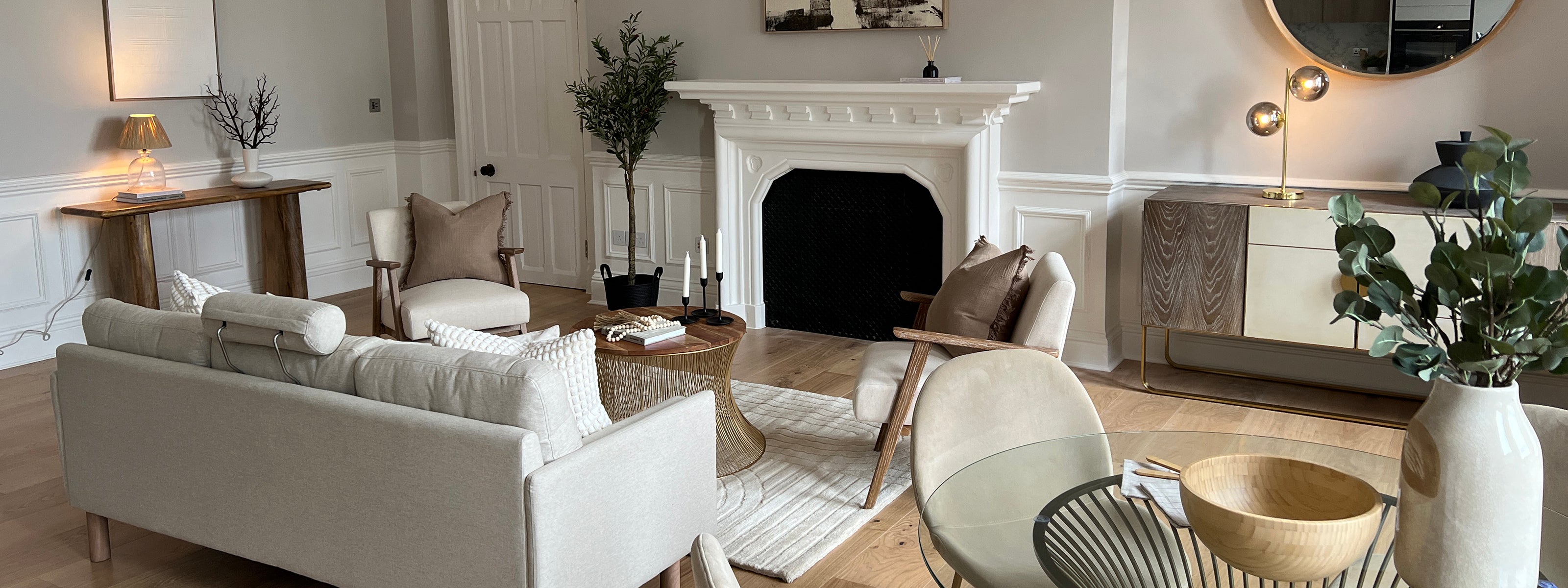 Stylish living room with a beige sofa, armchairs, white fireplace, round coffee table, and elegant decor featuring wood, plants, and soft lighting.