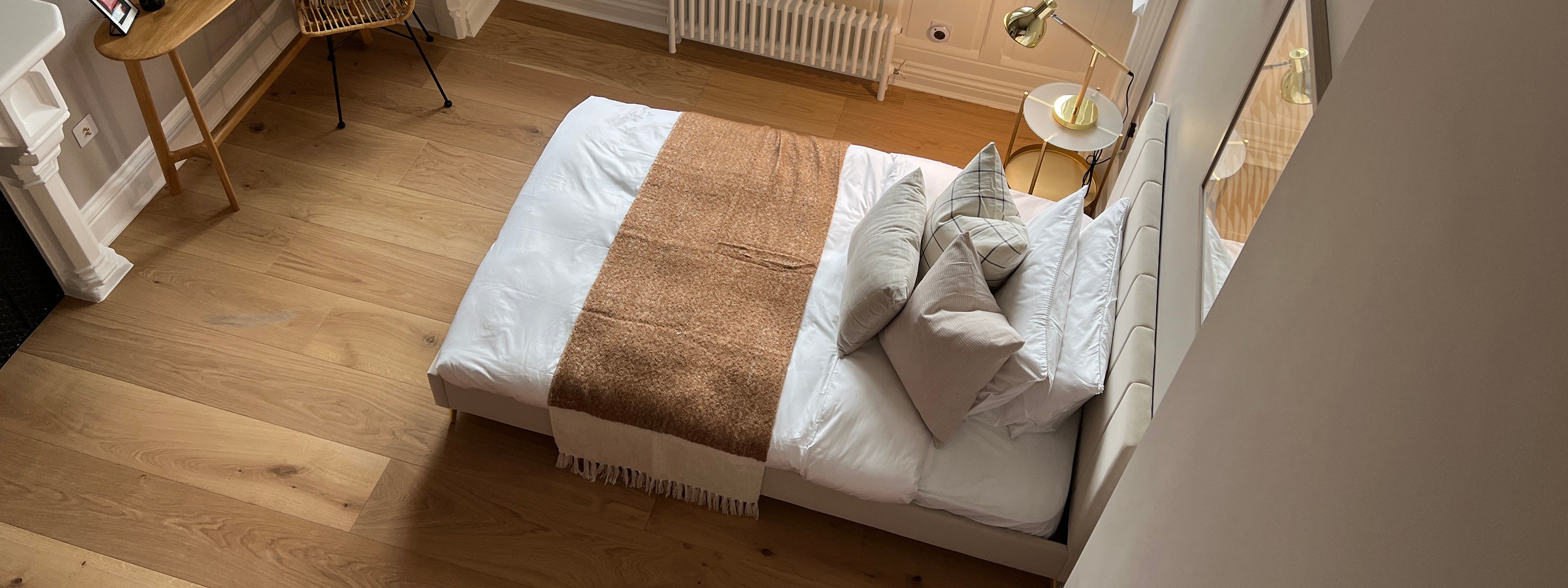 Top-down view of a modern bedroom featuring a neatly made bed with white linens, neutral-toned pillows, a brown throw blanket, wooden flooring, a minimalist nightstand with a gold lamp, and a small desk and chair in the corner.