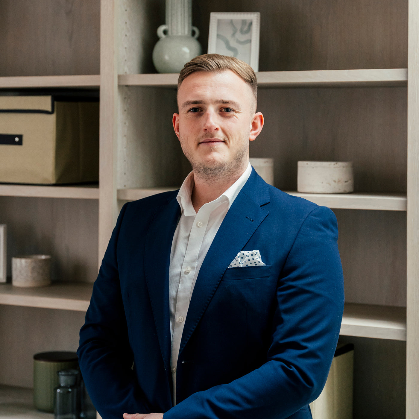 Young professional man in a tailored blue suit standing confidently in front of modern office shelving