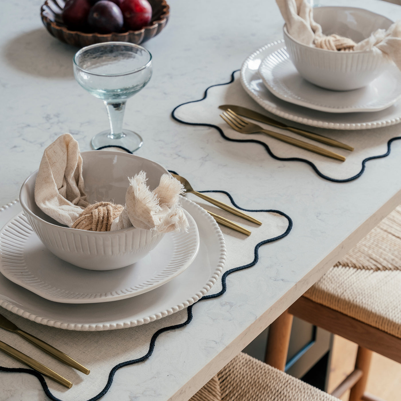 A close-up of an elegantly set dining table with a white marble surface. The place settings feature layered white ceramic plates and bowls, with beige napkins artistically arranged inside the bowls. Each setting includes gold-coloured cutlery placed on a beige placemat with a black wavy border. A textured woven stool and a glass goblet are also visible, with a bowl of plums in the background adding a touch of colour.