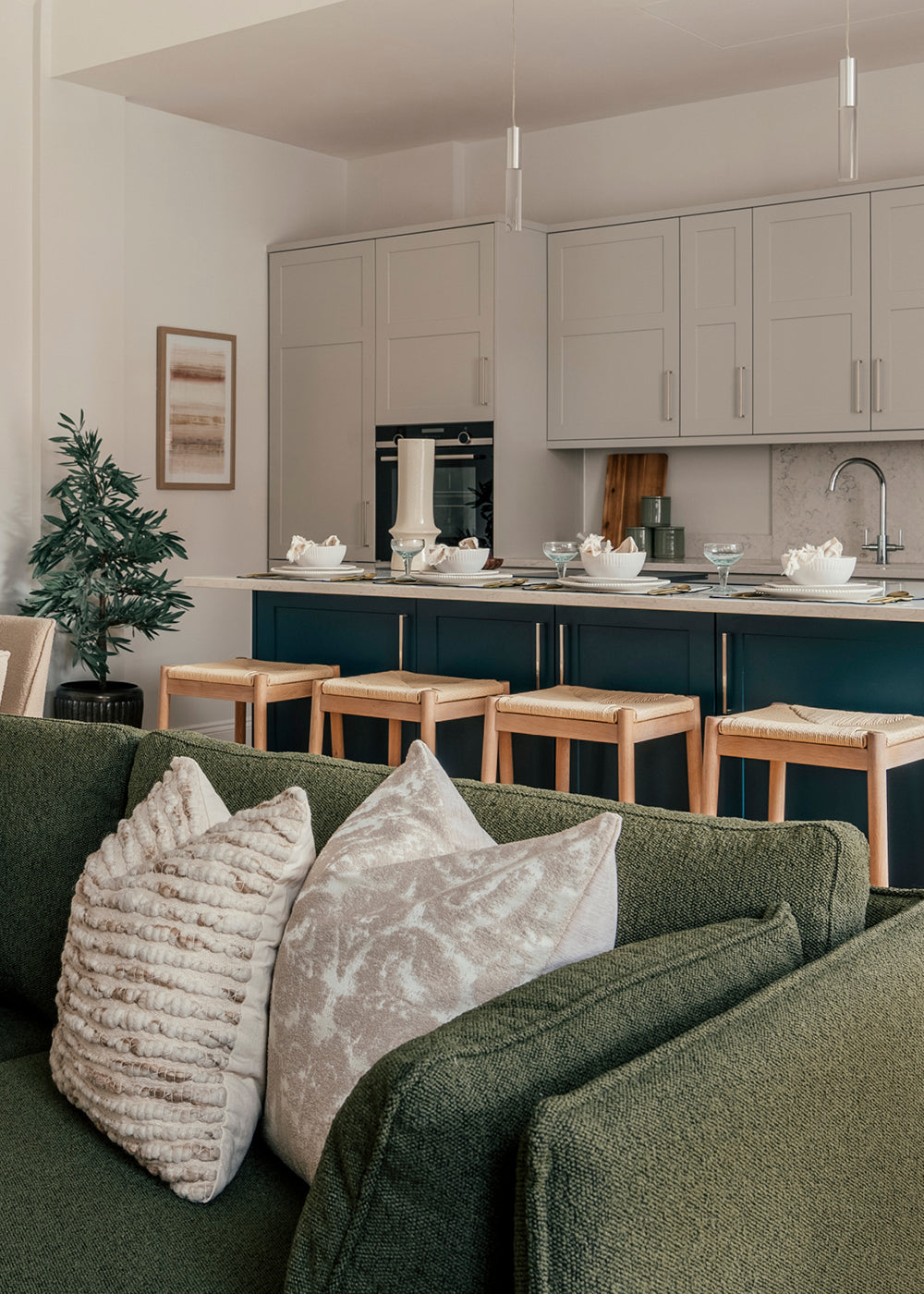 A modern open-plan kitchen and living space featuring a sage green sofa with textured neutral pillows in the foreground. The kitchen includes a large island with a white countertop, navy blue base, and four wooden bar stools. Above the island hang two minimalist pendant lights, and the back wall is lined with light grey cabinetry, a built-in oven, and a marble backsplash. A potted plant and framed artwork add natural and decorative touches to the space.
