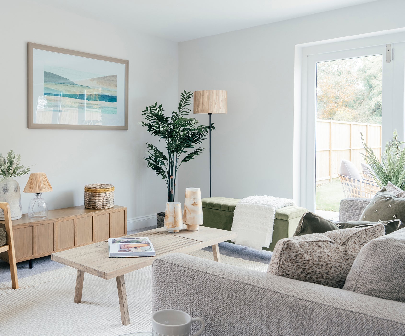 Bright and airy living room with a neutral color palette, featuring a light grey sofa with textured cushions, a wooden coffee table with a magazine, and a large window leading to a patio area with outdoor seating. A floor lamp and green plants add warmth and vibrancy to the space.