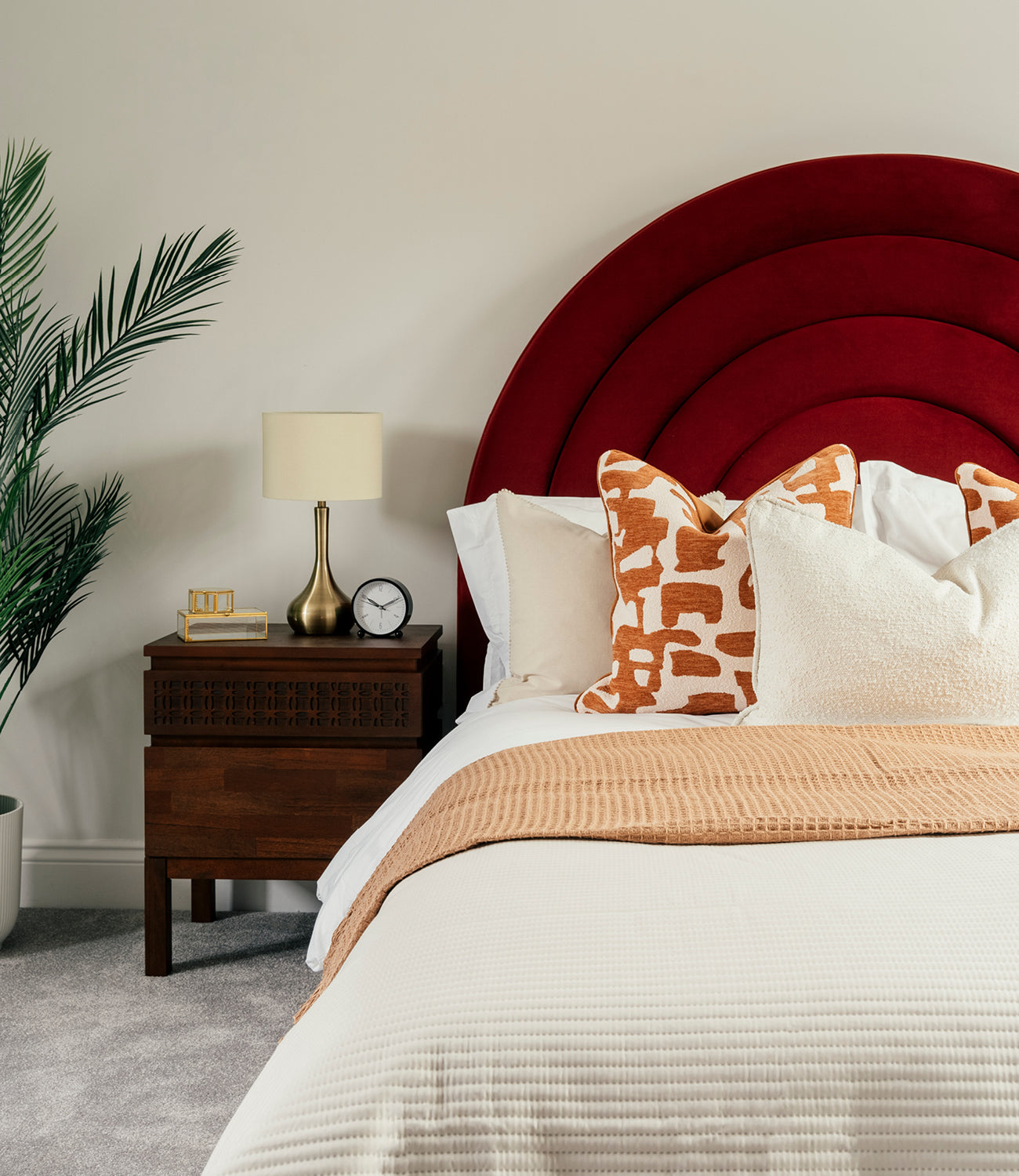 A stylish bedroom featuring a bold, curved red velvet headboard, a neatly made bed with white and beige bedding, accented by orange patterned and textured cream cushions, and a matching beige throw; a dark wooden nightstand holds a brass lamp, a small clock, and decorative items, with a potted palm adding greenery in the corner.