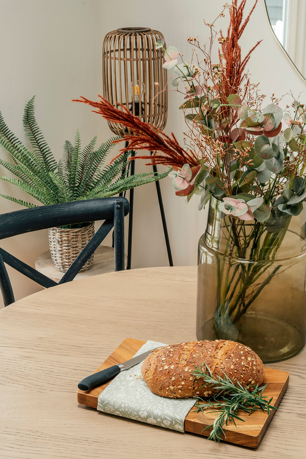 A close-up of a cozy dining corner featuring a round wooden table with a cutting board holding a loaf of seeded bread, a knife, a sprig of rosemary, and a green patterned cloth; in the background are a large glass vase with a dried floral arrangement, a wicker lamp, and a potted fern on a side table next to a black wooden chair.