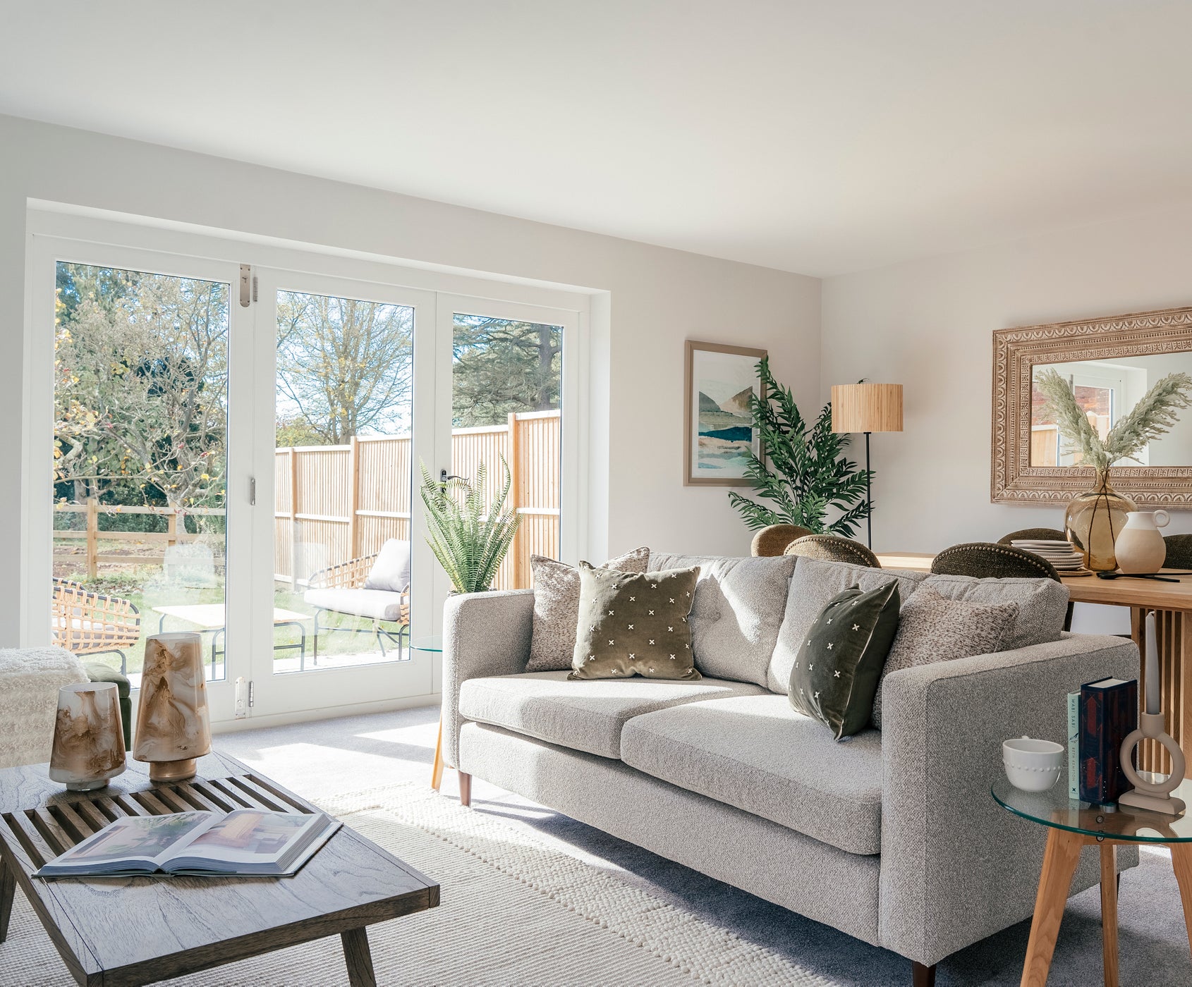 Bright and cozy living room featuring a light gray sofa with green and patterned cushions, a wooden coffee table with decorative vases and an open magazine, and large glass doors opening to a garden patio with outdoor seating; neutral tones and natural textures create a welcoming atmosphere.