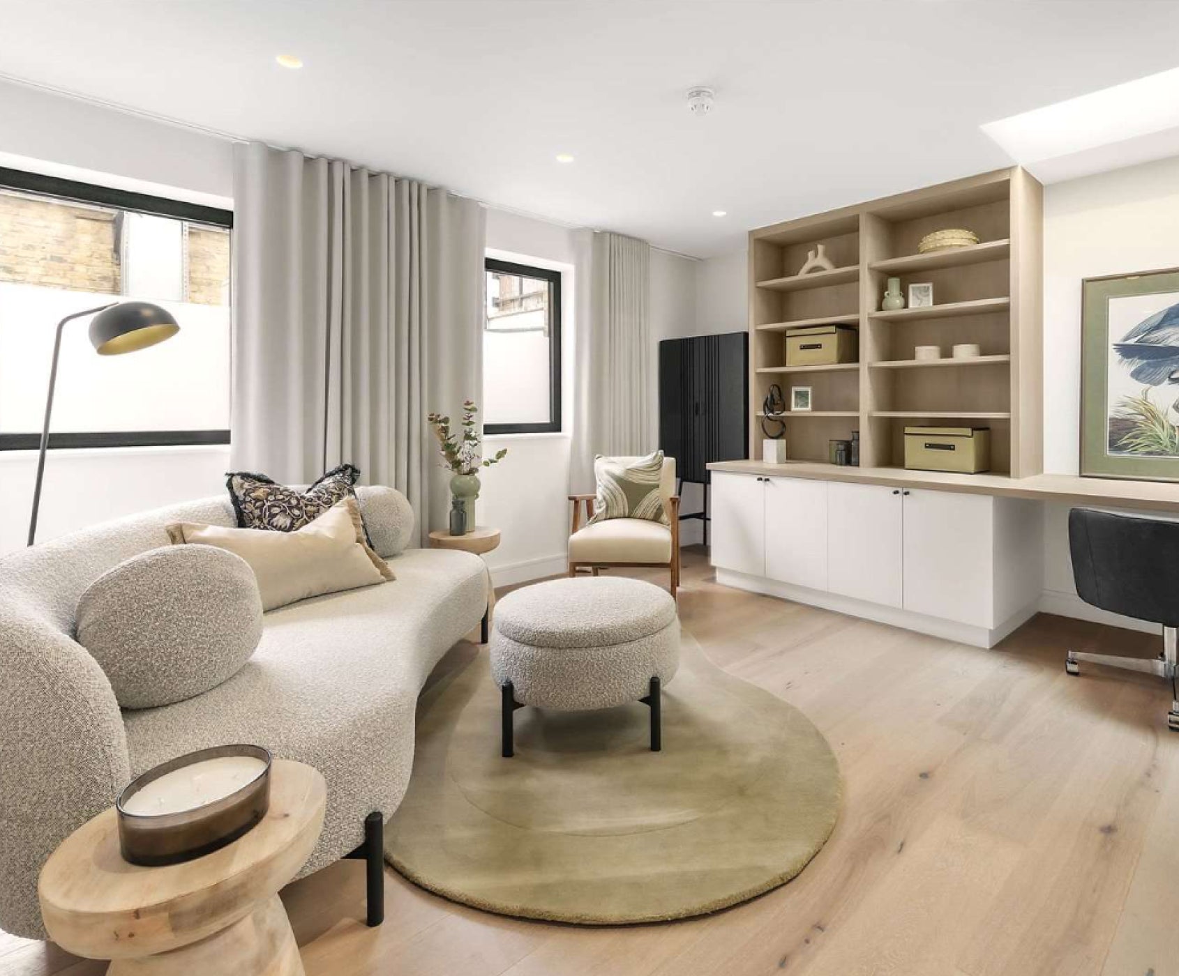 Modern home office and living area featuring a curved boucle sofa, neutral drapes, built-in shelving, natural wood flooring, and minimalist decor.