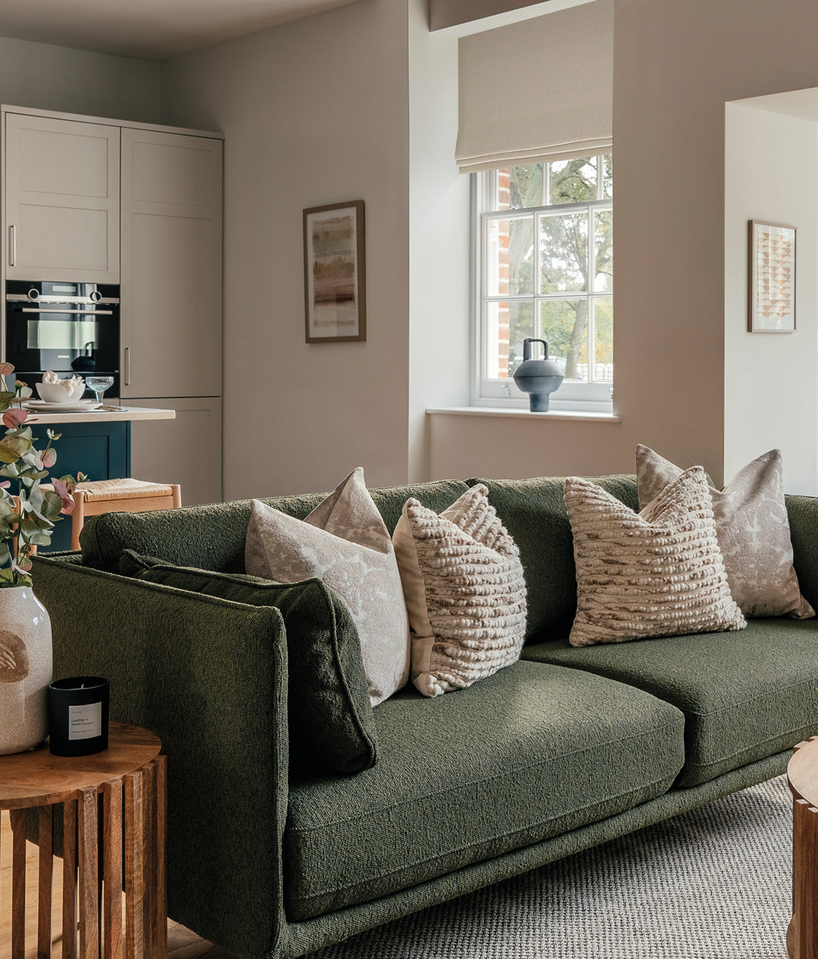 Stylish living room featuring a textured green sofa with neutral cushions, wooden side tables, modern decor, and a kitchen in the background with light cabinetry.