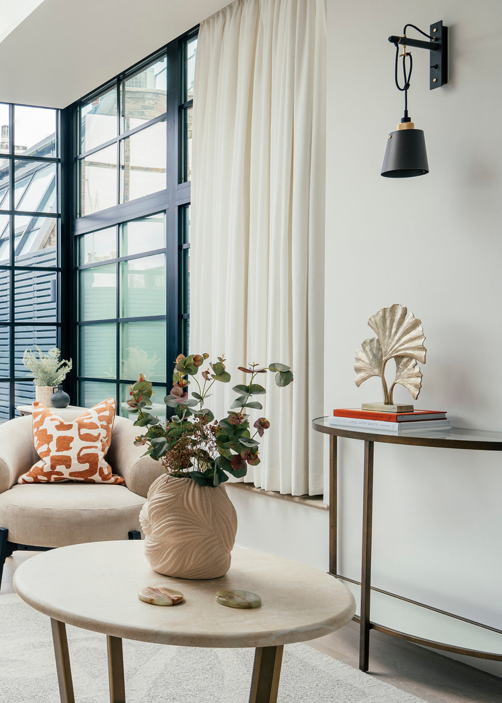 Modern living room with large industrial-style windows, cream curtains, a cozy armchair with a patterned cushion, and a round coffee table with a decorative vase and greenery.